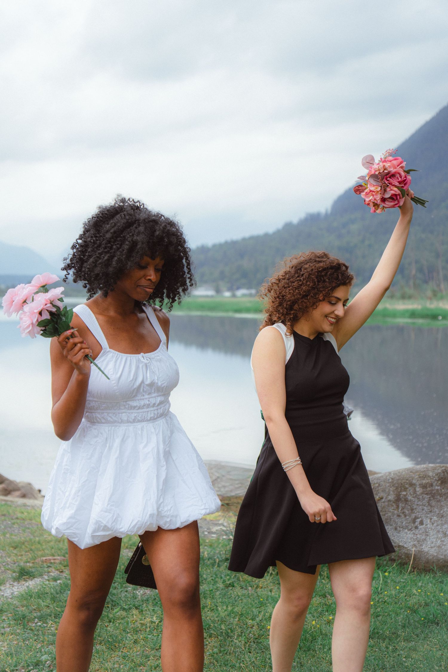 Two women in black and white dresses holding pink flower bouquets near a mountain lake.
