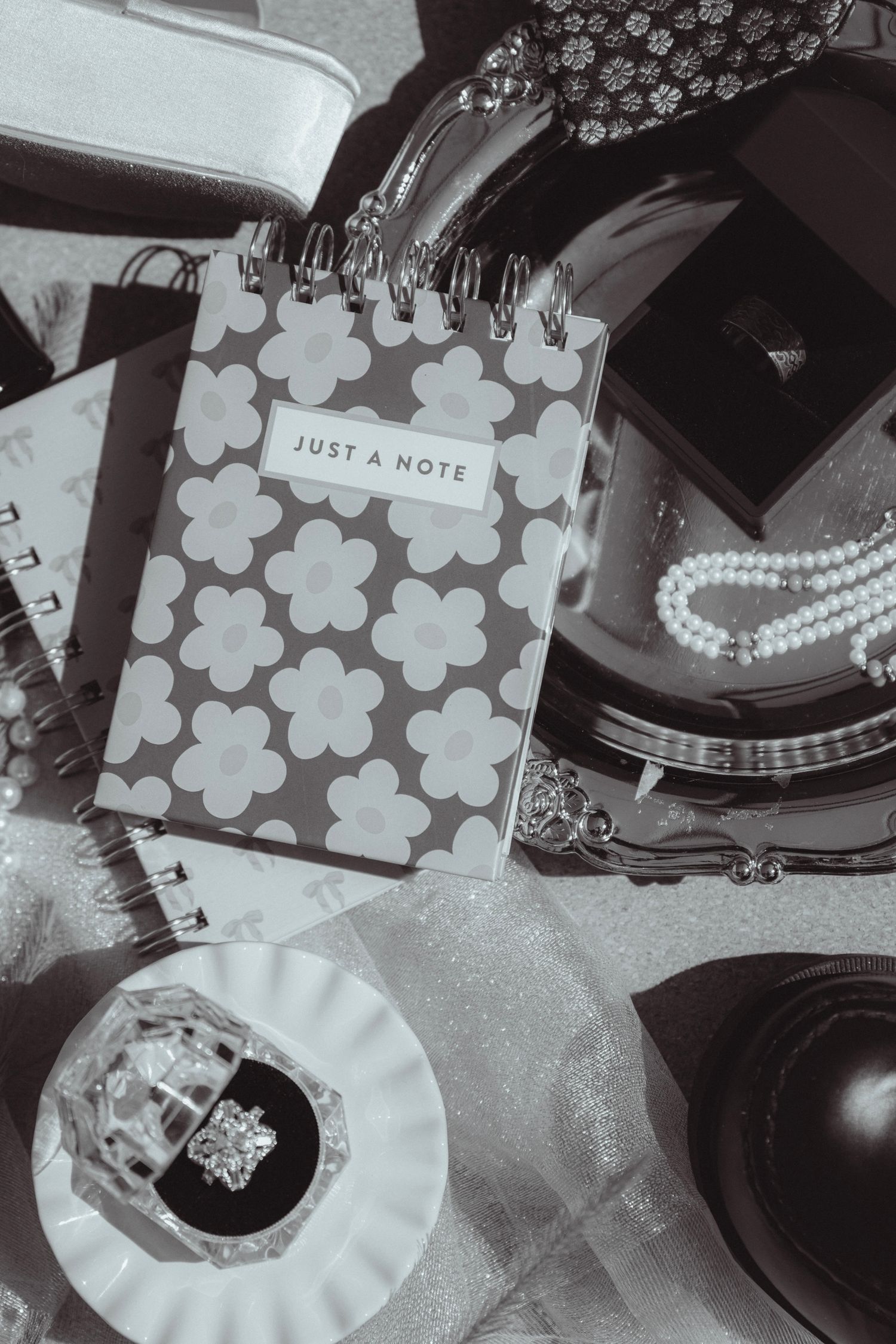 Black and white flatlay of a floral notepad, pearl necklace, ring in dish, and elegant accessories on a table.