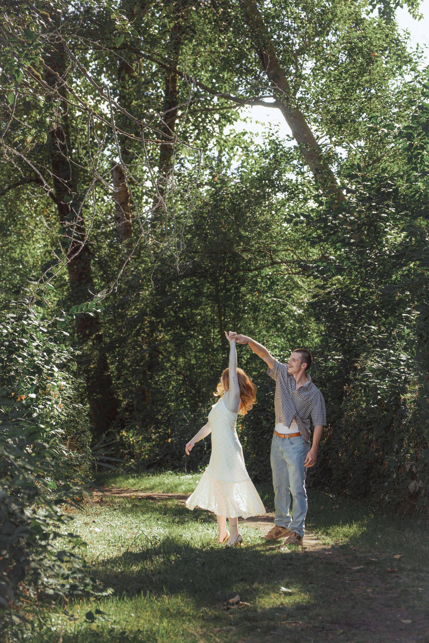 Couple dancing together in a lush green forest path, woman in white dress twirling under man's raised arm.
