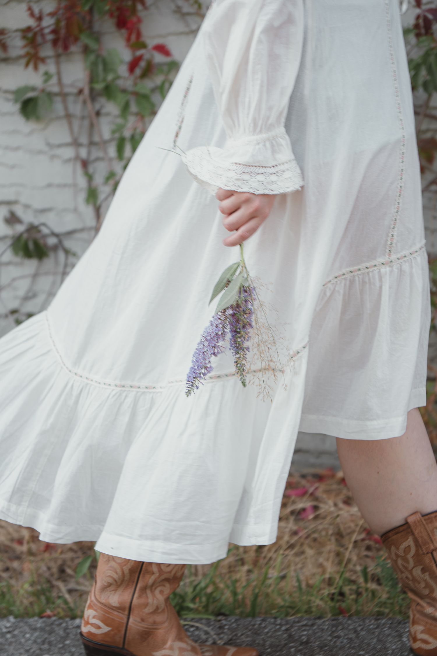 Close-up of white tiered lace dress with brown cowboy boots holding purple lavender bouquet outdoors.