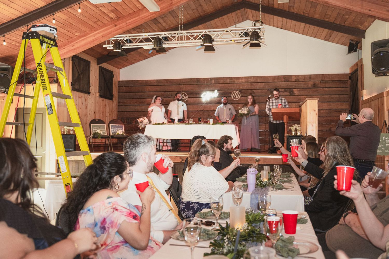 Guests seated at long tables in a rustic barn venue watch a presentation at the front of the room.