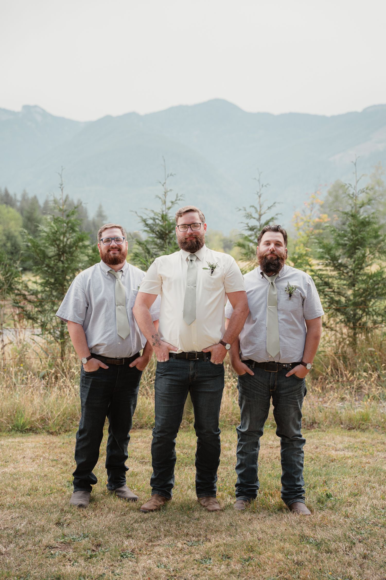 Three bearded groomsmen in ties and jeans pose outdoors with mountain and pine tree backdrop.