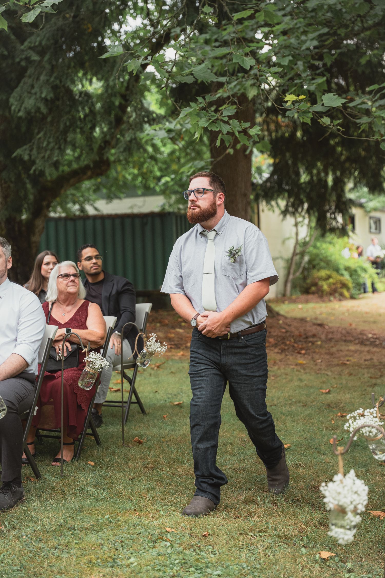 Groom in gray shirt with boutonniere awaiting bride at outdoor wedding ceremony.