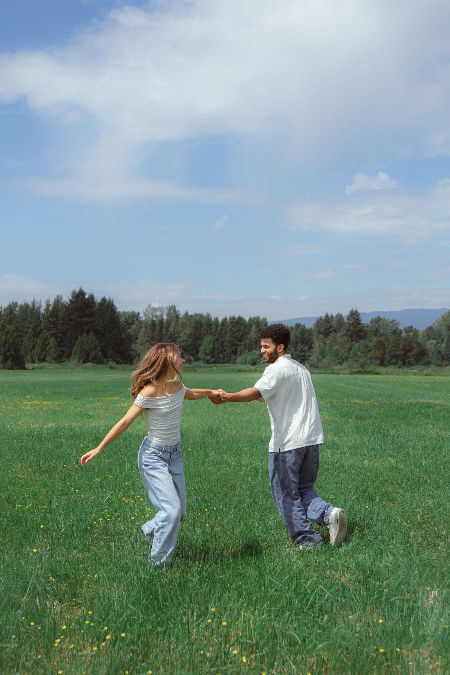 A playful couple holding hands and spinning together in a wide open green field with trees in the background.