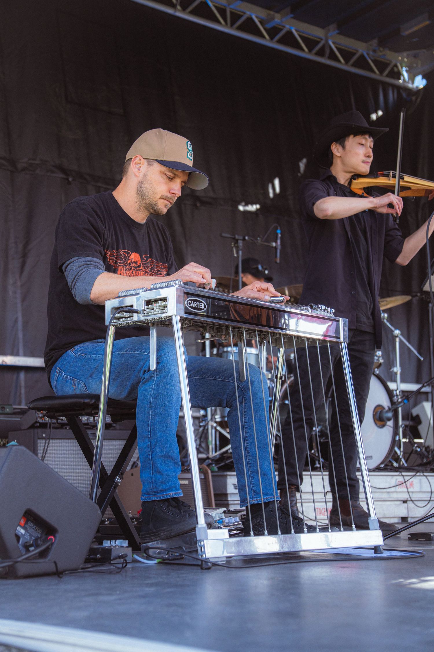 Musician playing pedal steel guitar on outdoor stage with violinist performing in background.