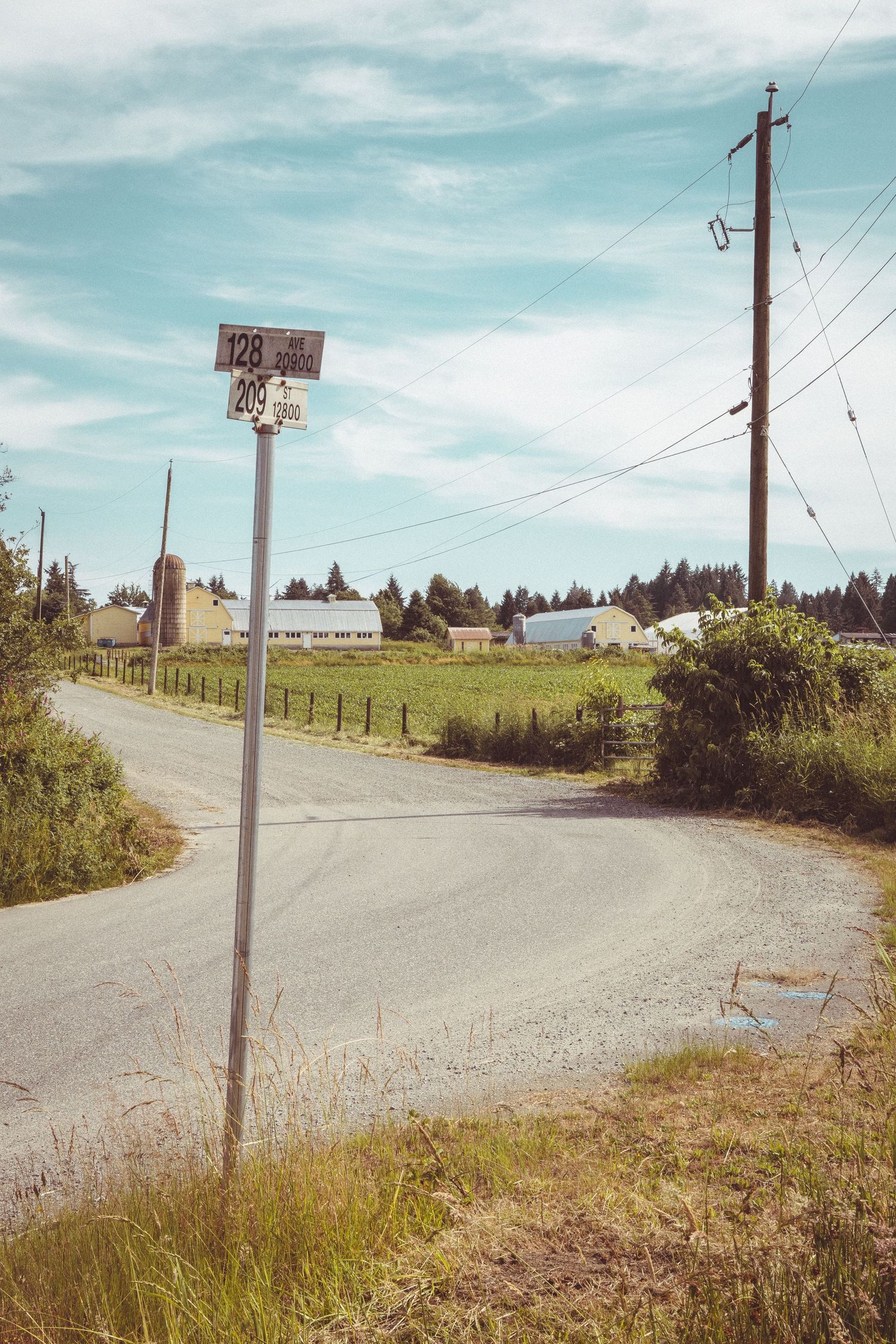 Rural road intersection with street signs, power lines, and farm buildings visible in distance.