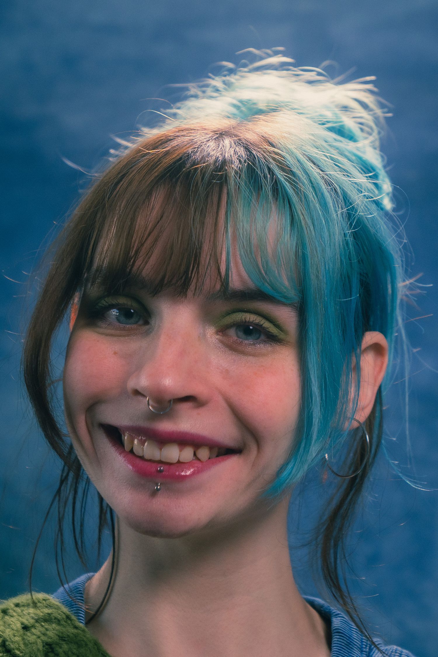 Smiling young woman with blue and brown hair, nose ring, and green eyeshadow in portrait photo.