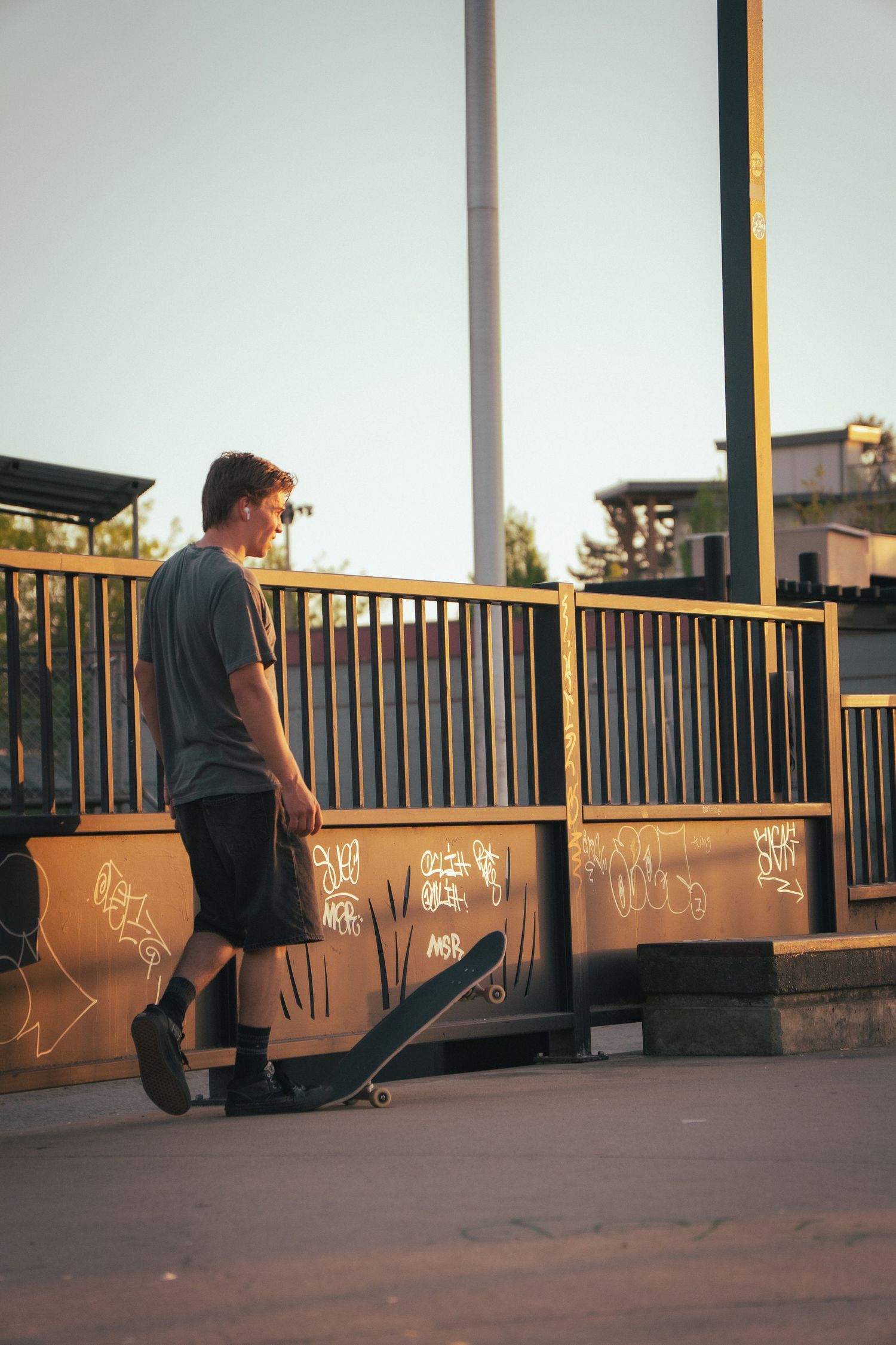 Skateboarder in grey shirt walking with a skateboard along a graffiti-covered fence at sunset.