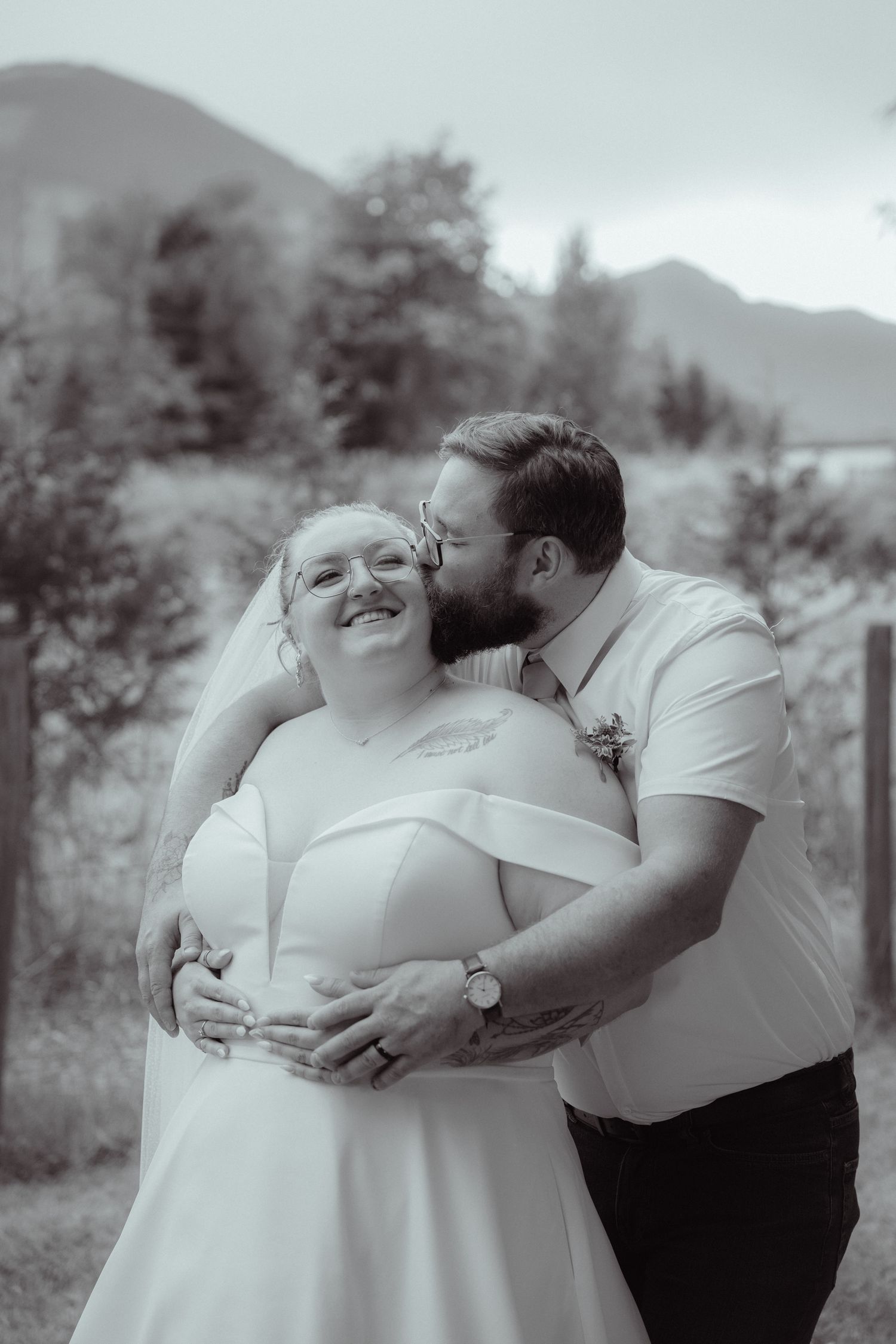 Black and white photo of a groom kissing his smiling bride on the cheek, embracing outdoors with mountains behind.