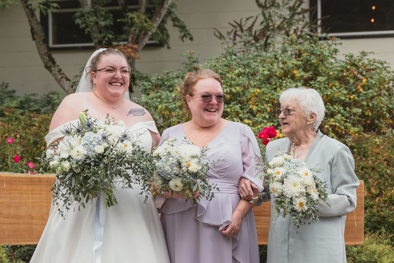 Bride and two women smiling outdoors, holding white floral bouquets with greenery at a wedding celebration.