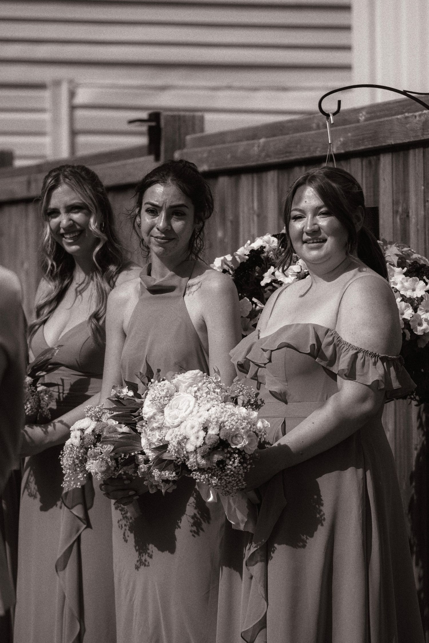 Three bridesmaids in matching gowns holding floral bouquets pose together outdoors in black and white wedding photo.
