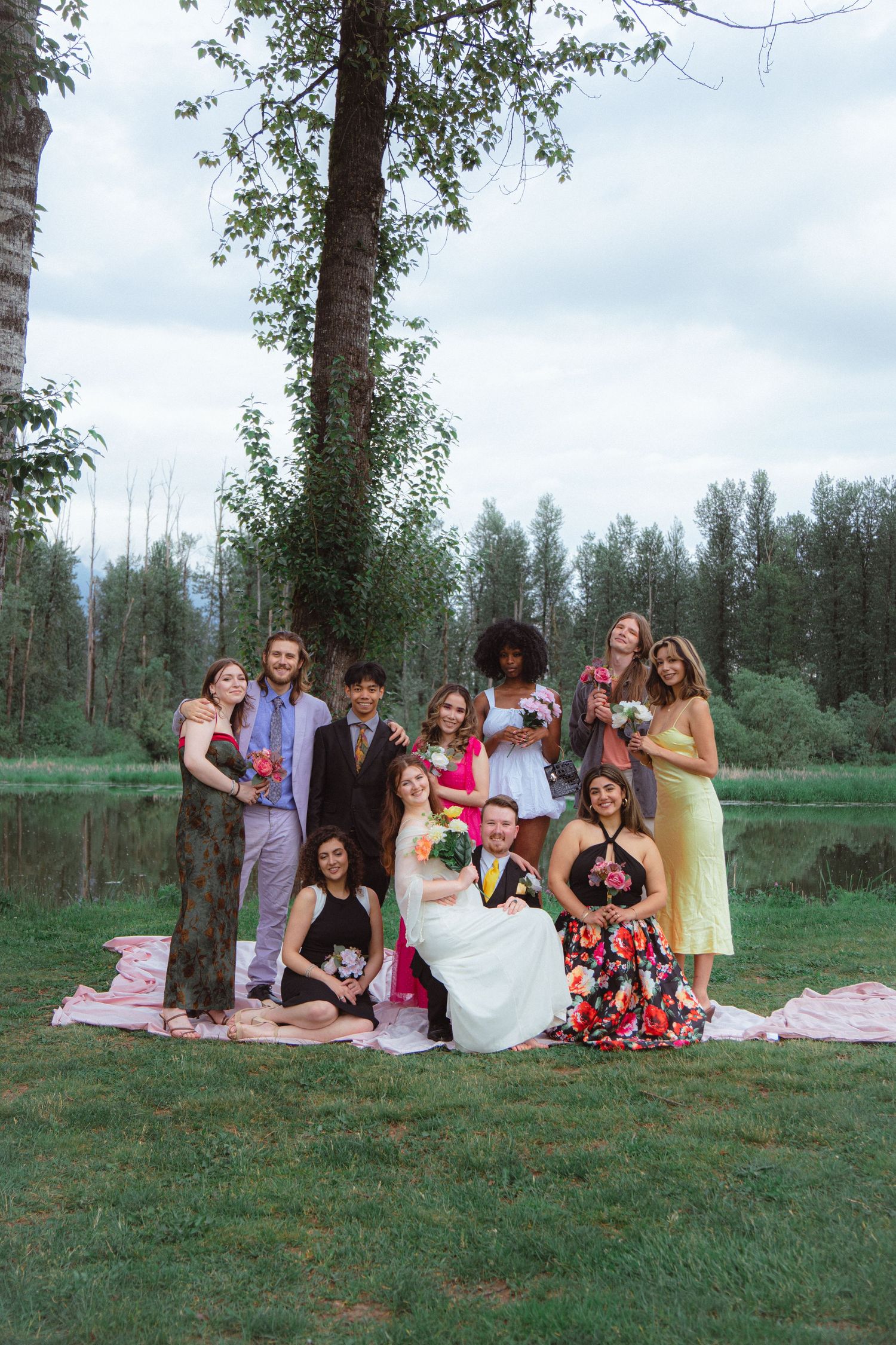 Wedding party posing outdoors by a lake in Mission British Columbia Canada, bride in white, guests in colourful attire, surrounded by trees.