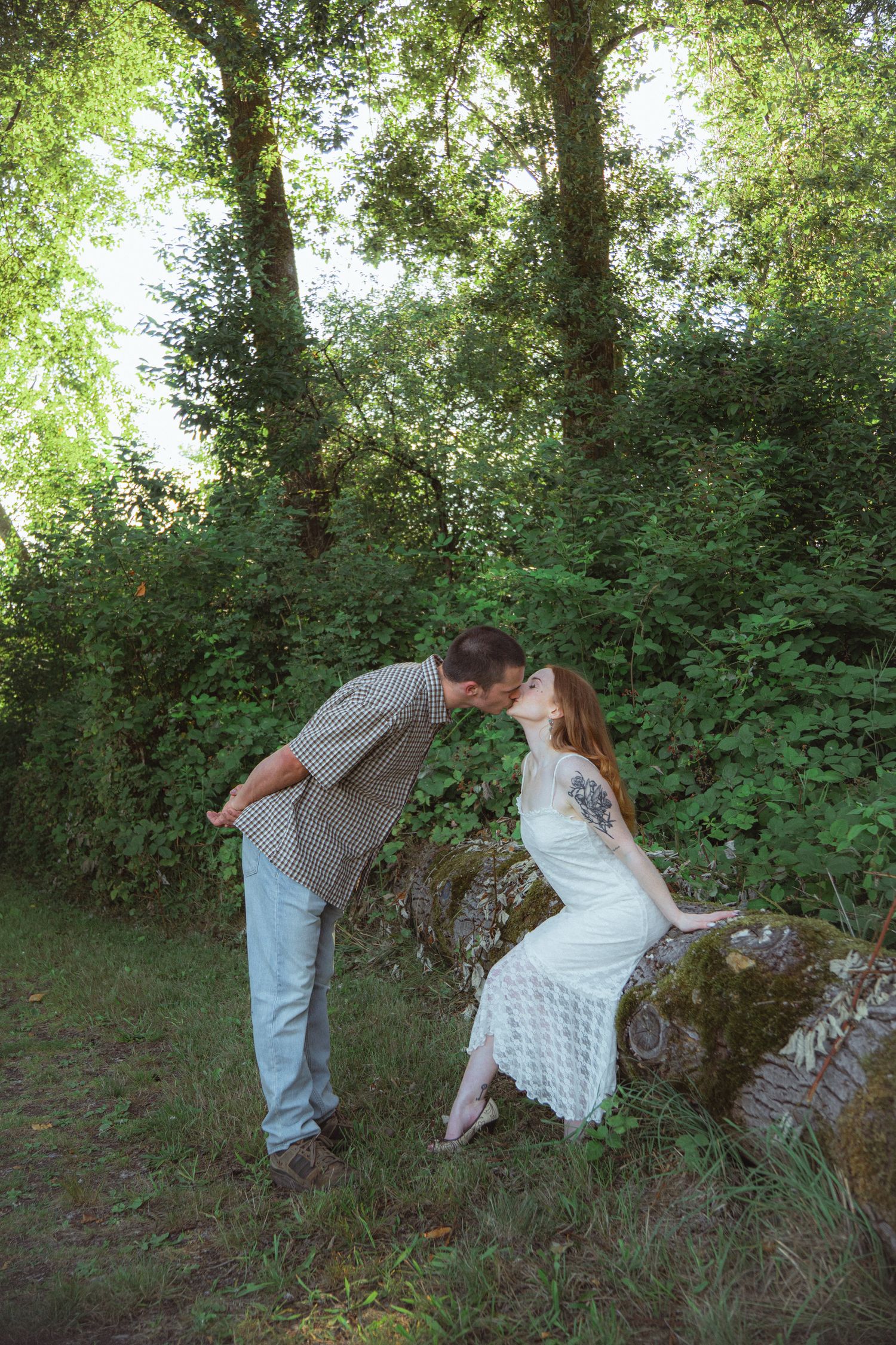 Couple sharing a romantic kiss outdoors, woman in white lace dress seated on mossy log, man in plaid shirt, lush green forest.