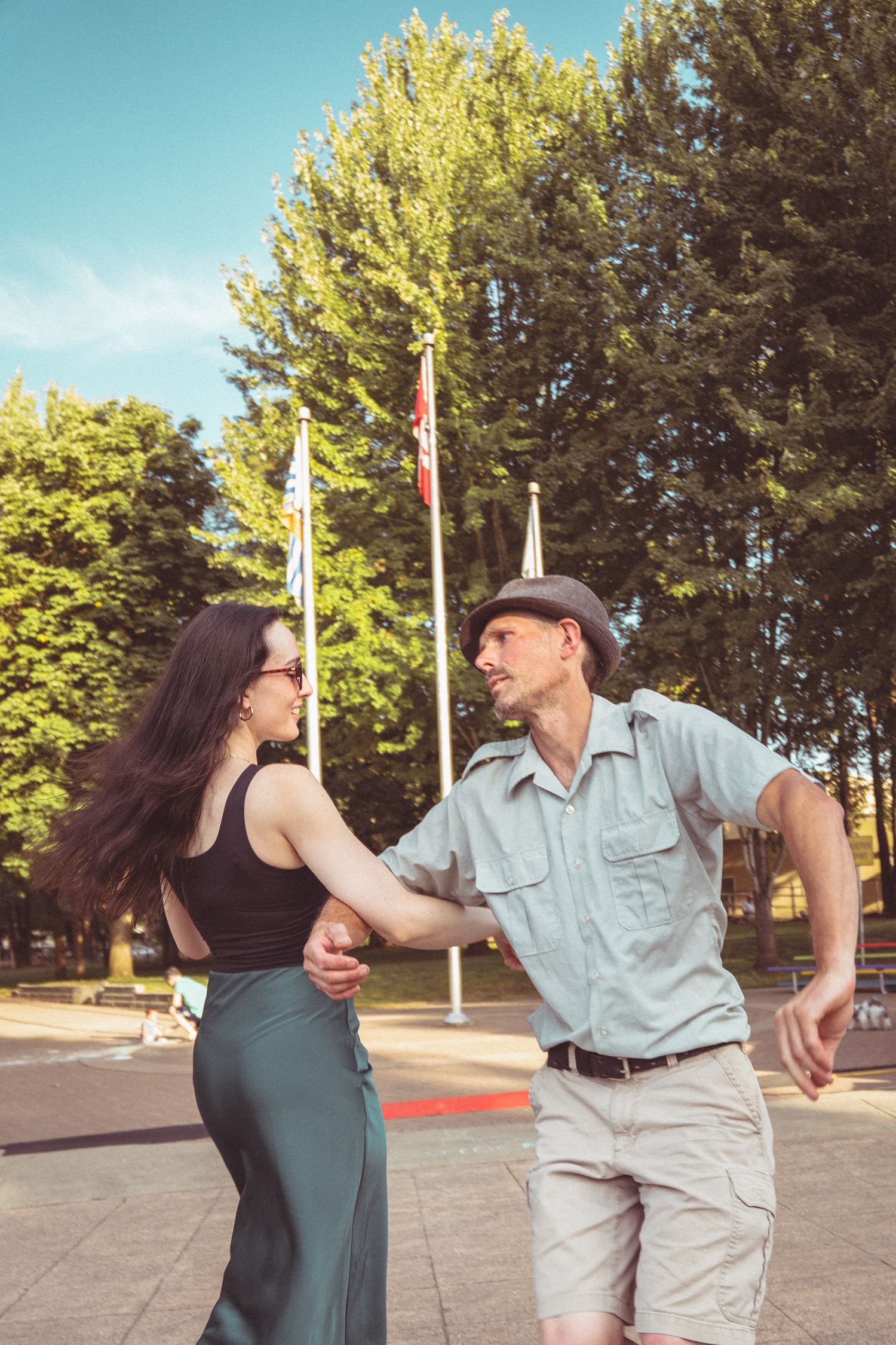 A couple dances outdoors in a sunny park, holding hands near flagpoles and lush green trees.