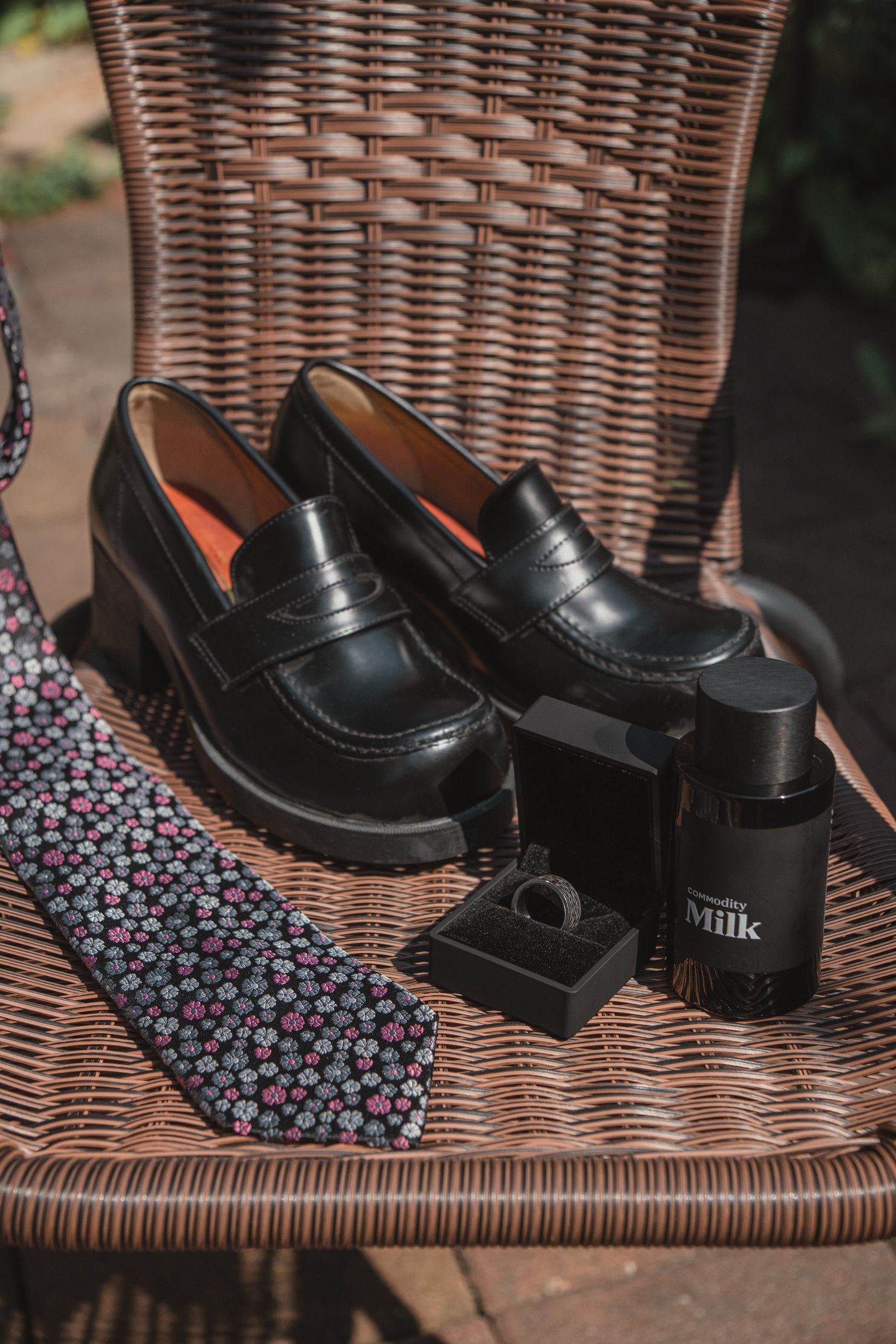 Black penny loafers, floral tie, ring, and leather milk conditioner arranged on a wicker chair for a wedding preparation.