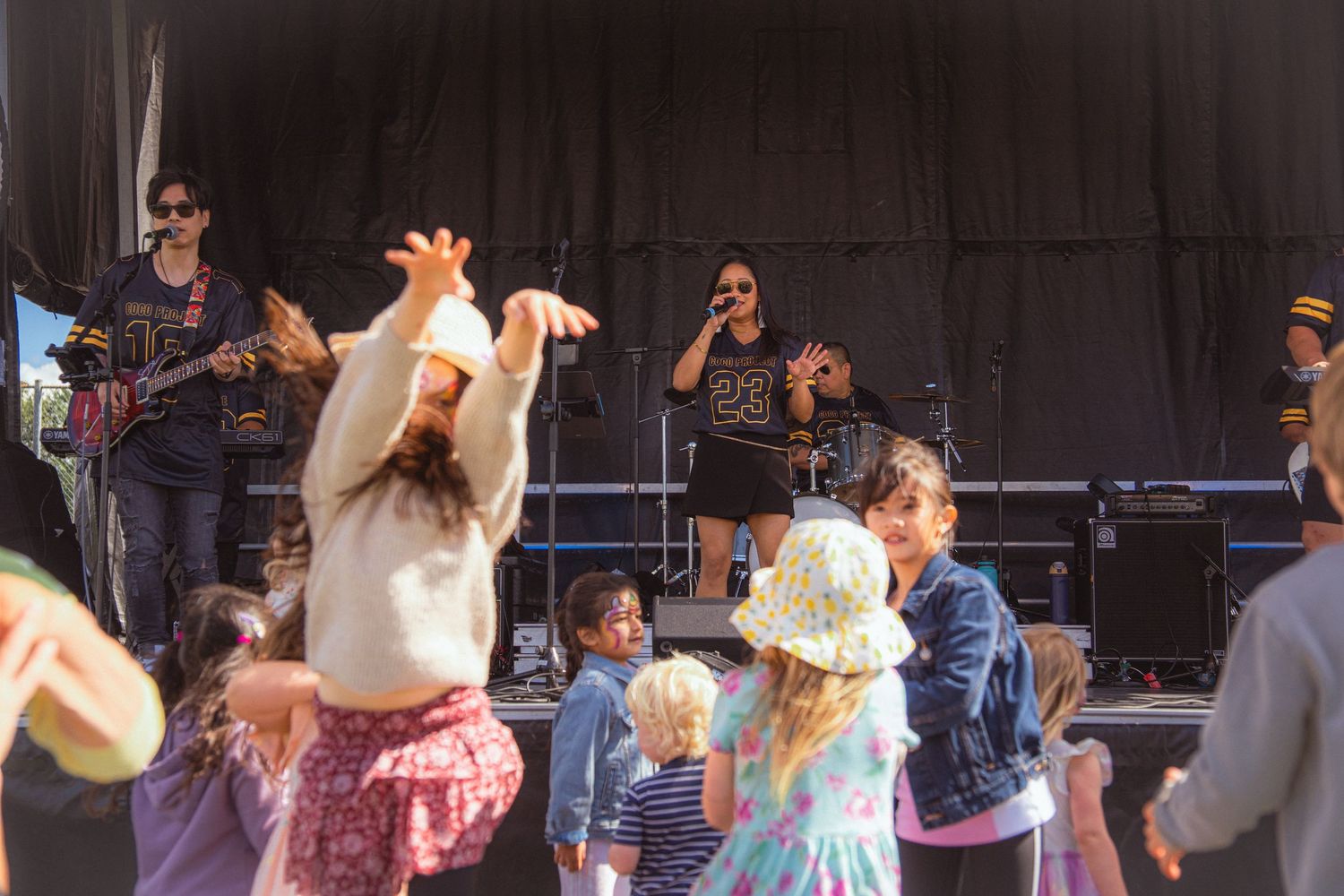 Band performs on outdoor stage as children dance and play in front of the crowd.