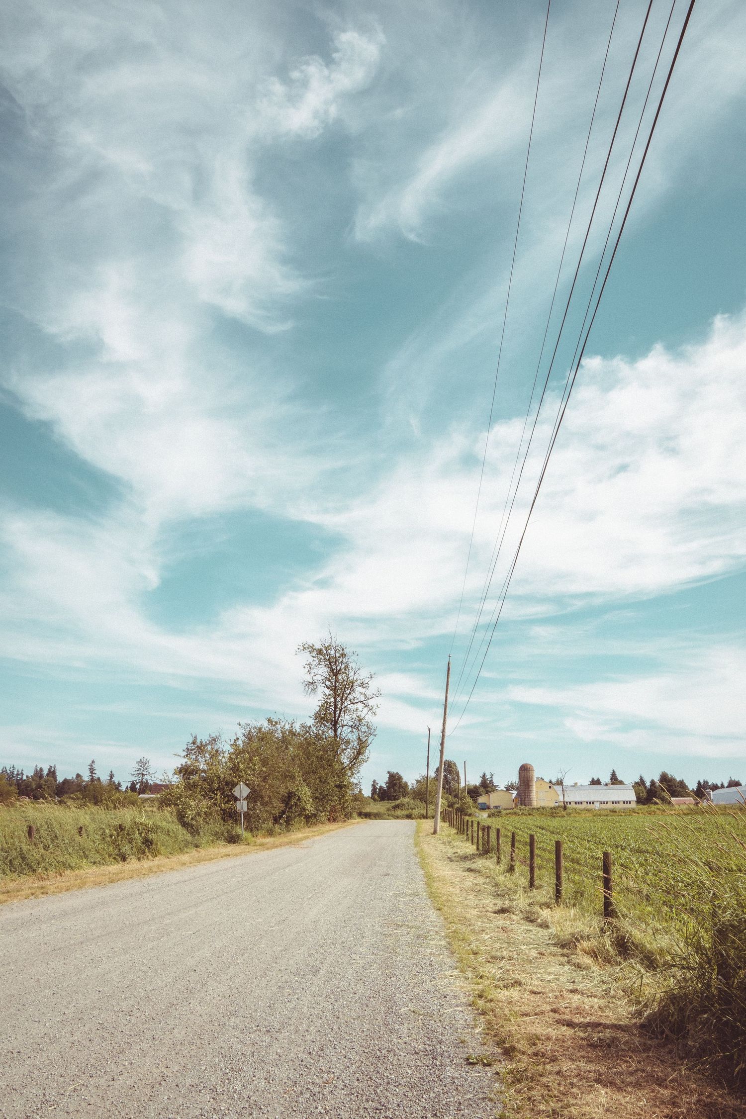 Quiet country road with power lines, wooden fence, green fields and farm buildings under a cloudy blue sky.