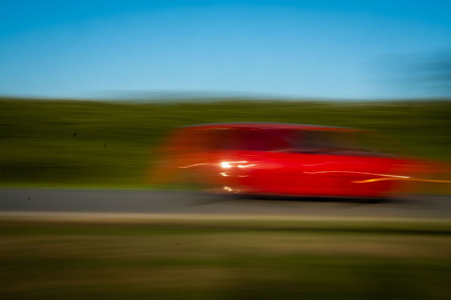 A red car captured with motion blur speeding along a road under a clear blue sky.