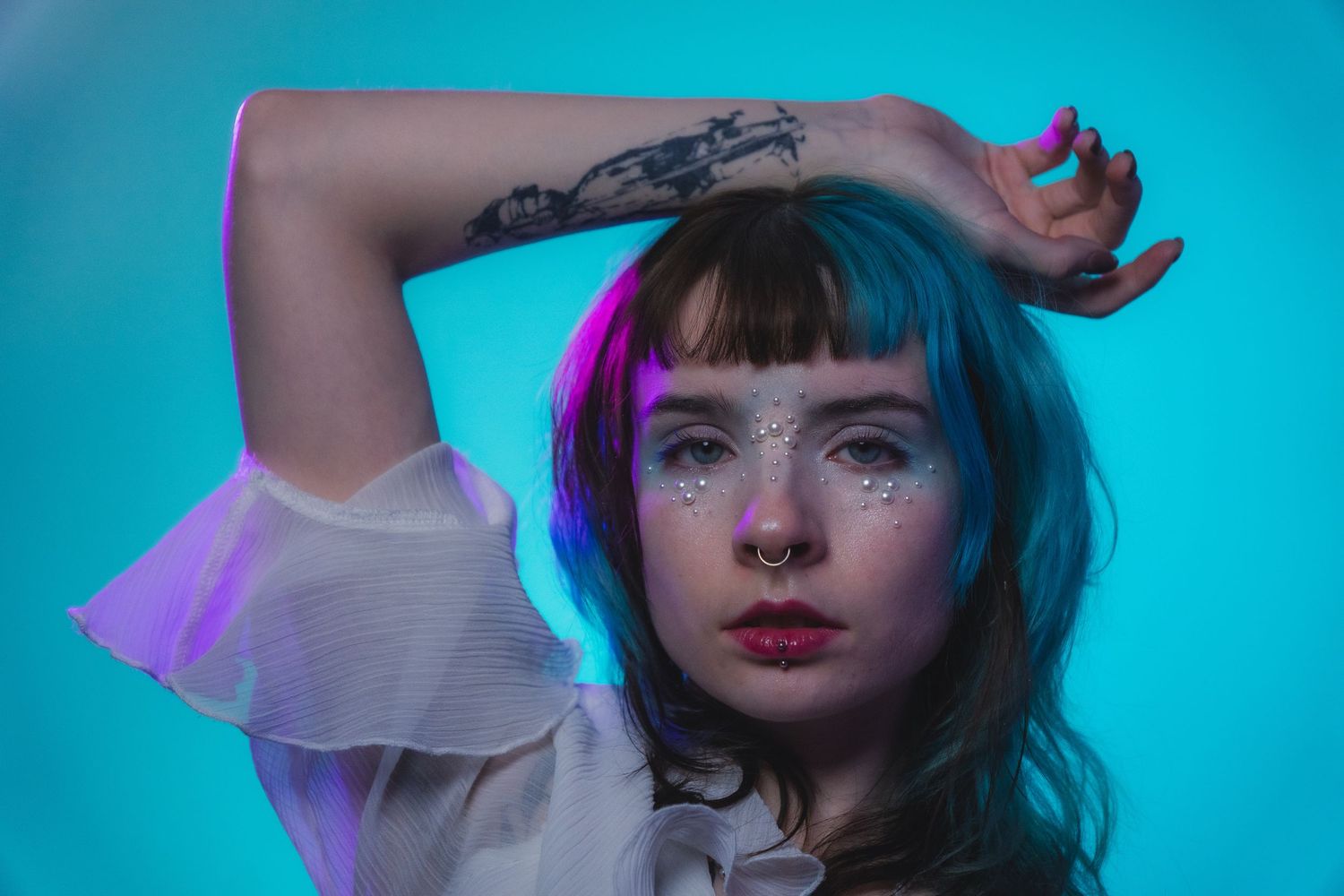 Woman with blue hair and arm tattoo posing against a teal background with glitter face gems.