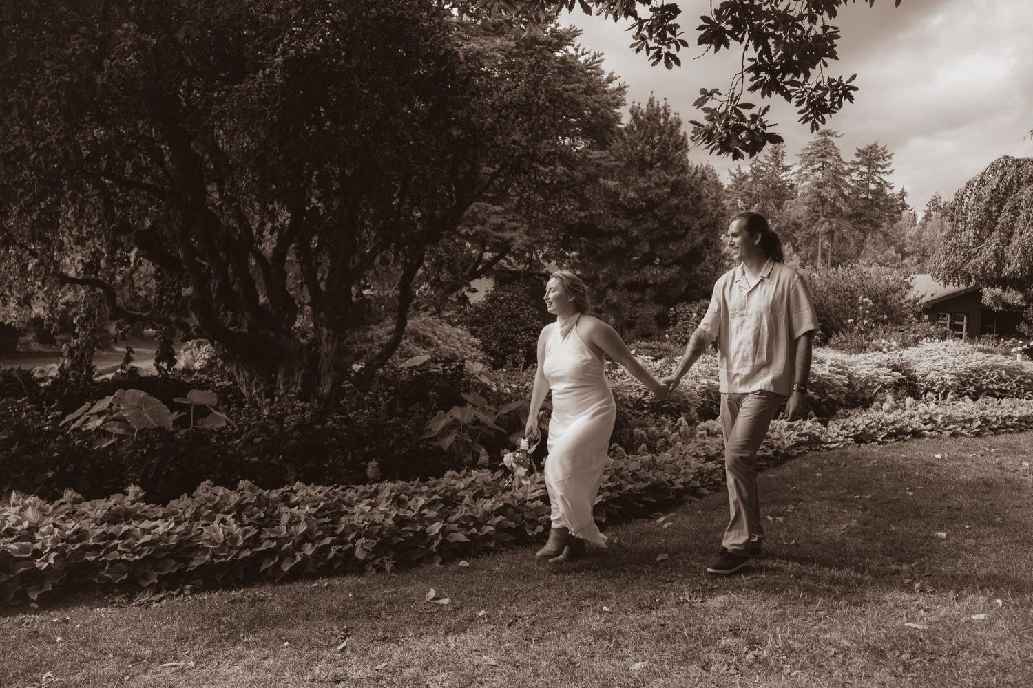 Couple holding hands walking through lush garden in black and white photo.