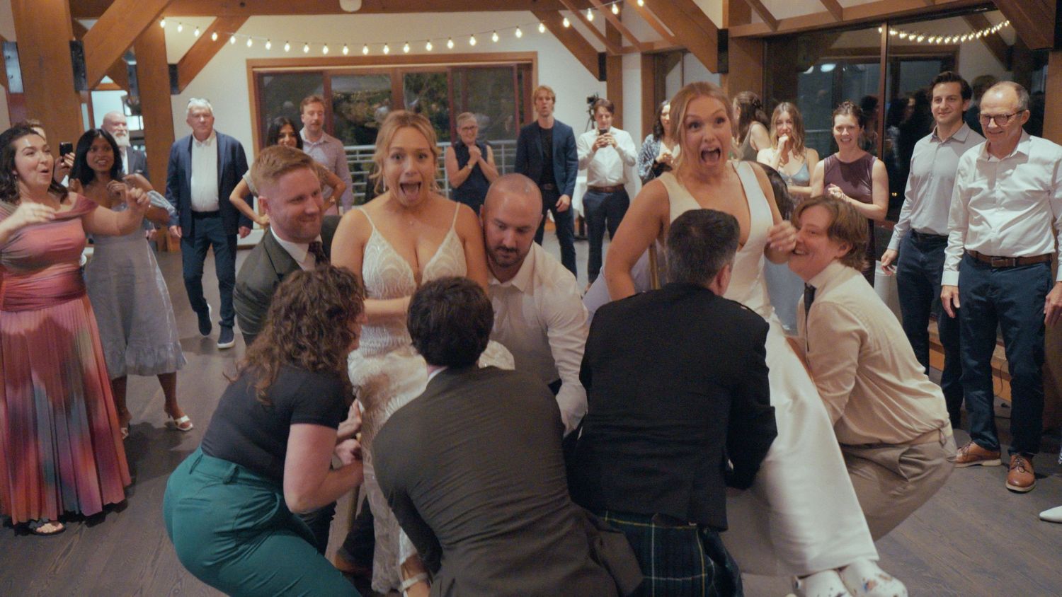 Wedding guests gather around bride and groom on dance floor during lively reception celebration indoors.