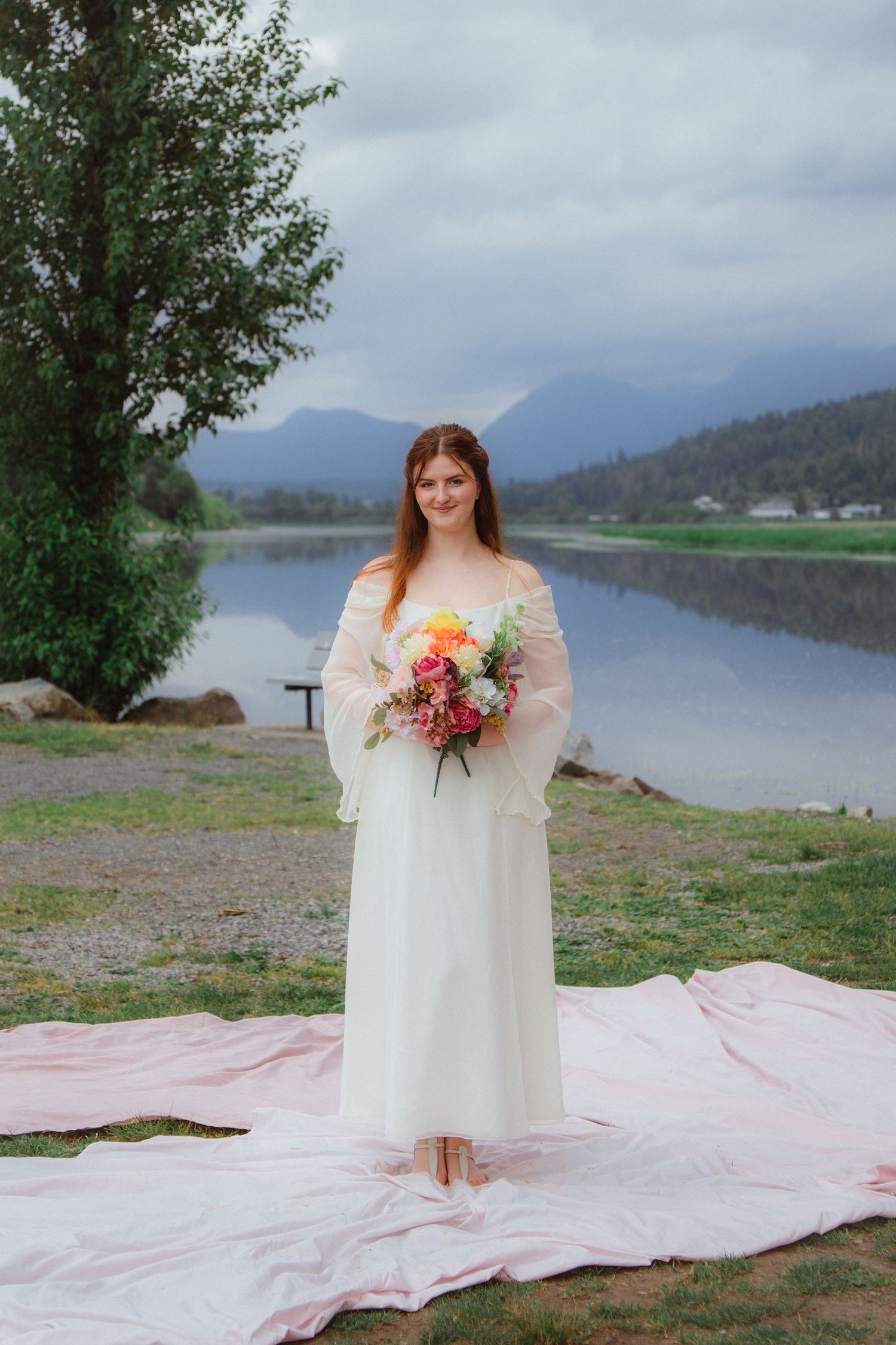 Bride in white off-shoulder gown holds colorful bouquet by a serene mountain lake.