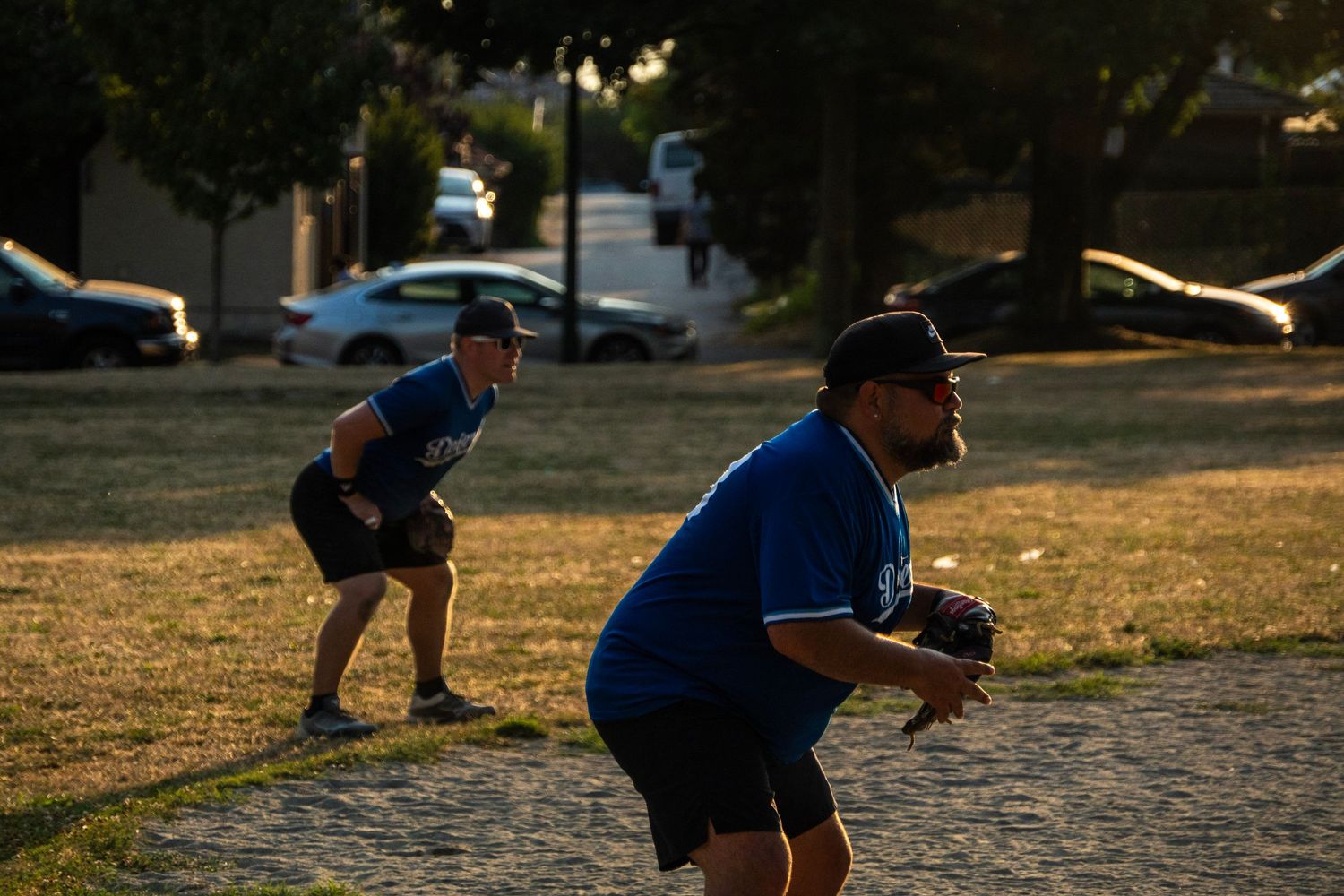 Two softball players in blue uniforms fielding positions on a sunlit grass field at golden hour.