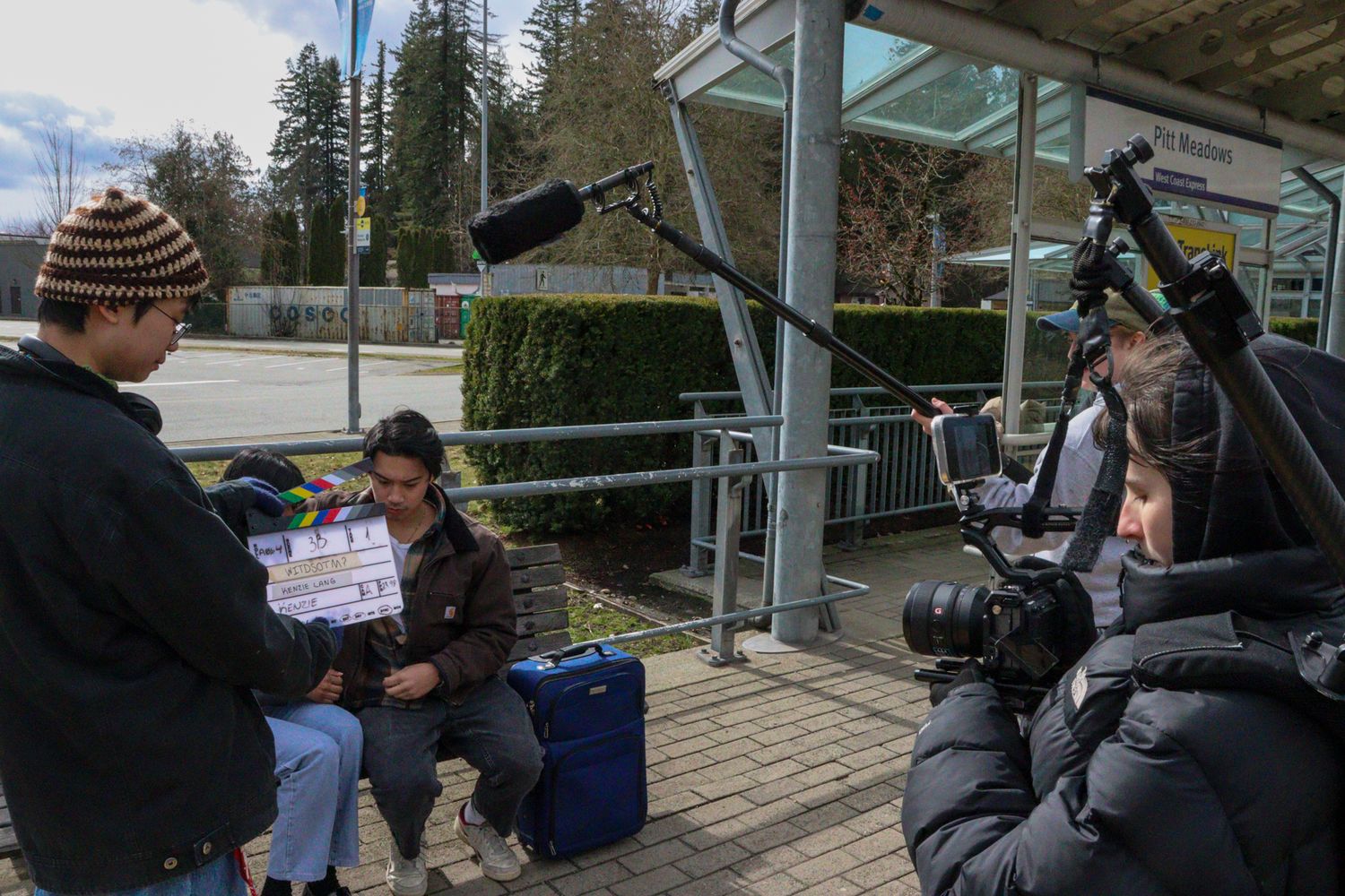 Film crew shooting a scene with actor at an outdoor bus stop location.