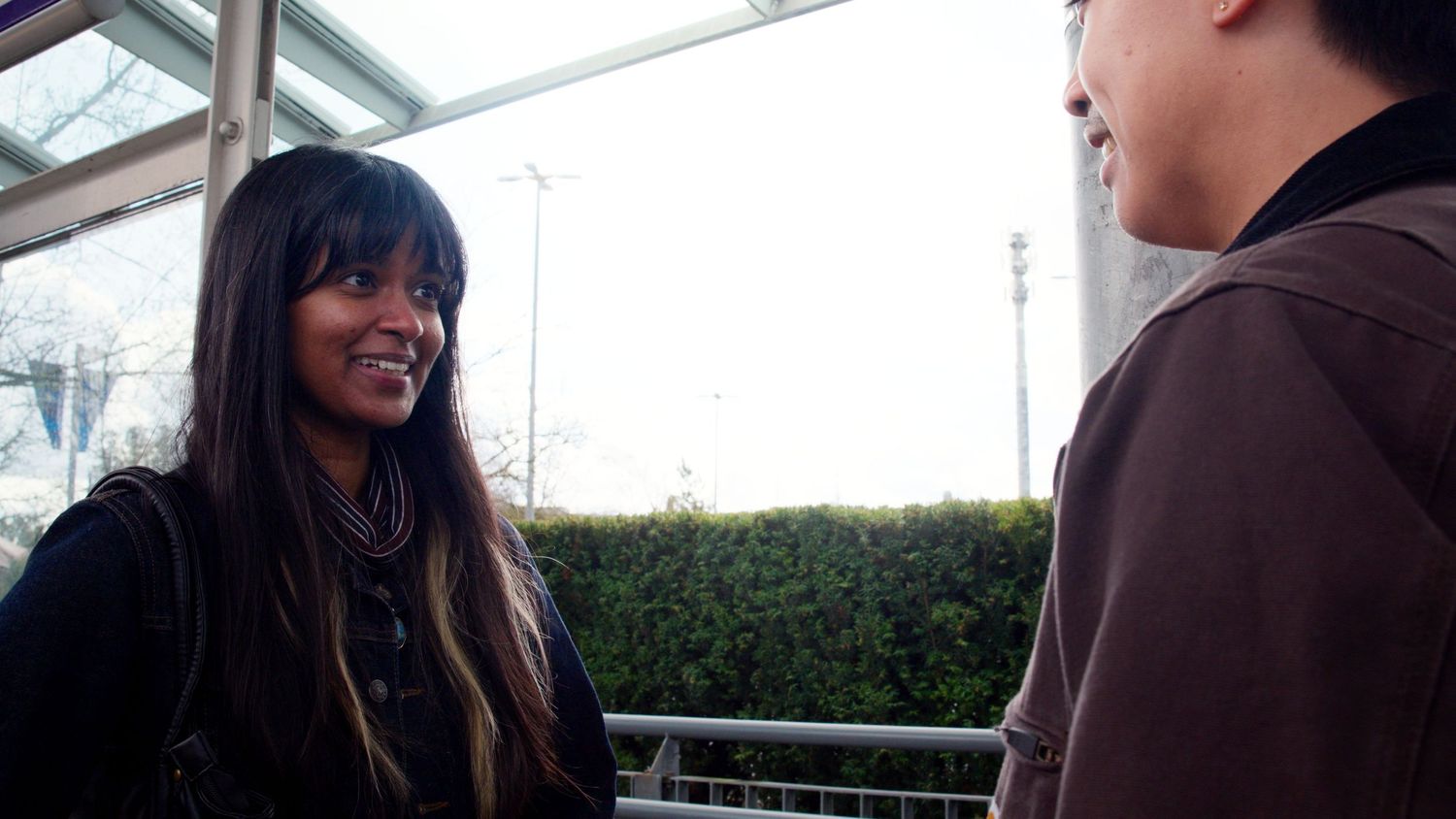 Young woman with long hair smiles warmly at someone outside a transit station on a sunny day.