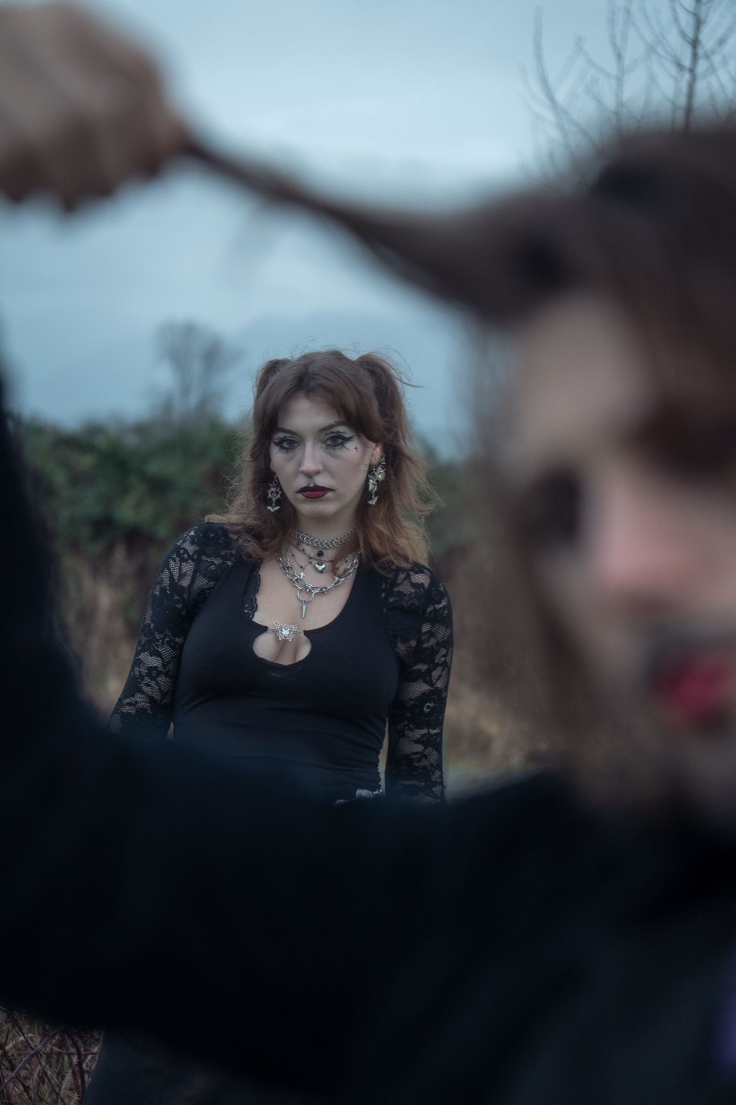 A gothic woman in black lace dress with silver necklace poses outdoors on a cloudy day.
