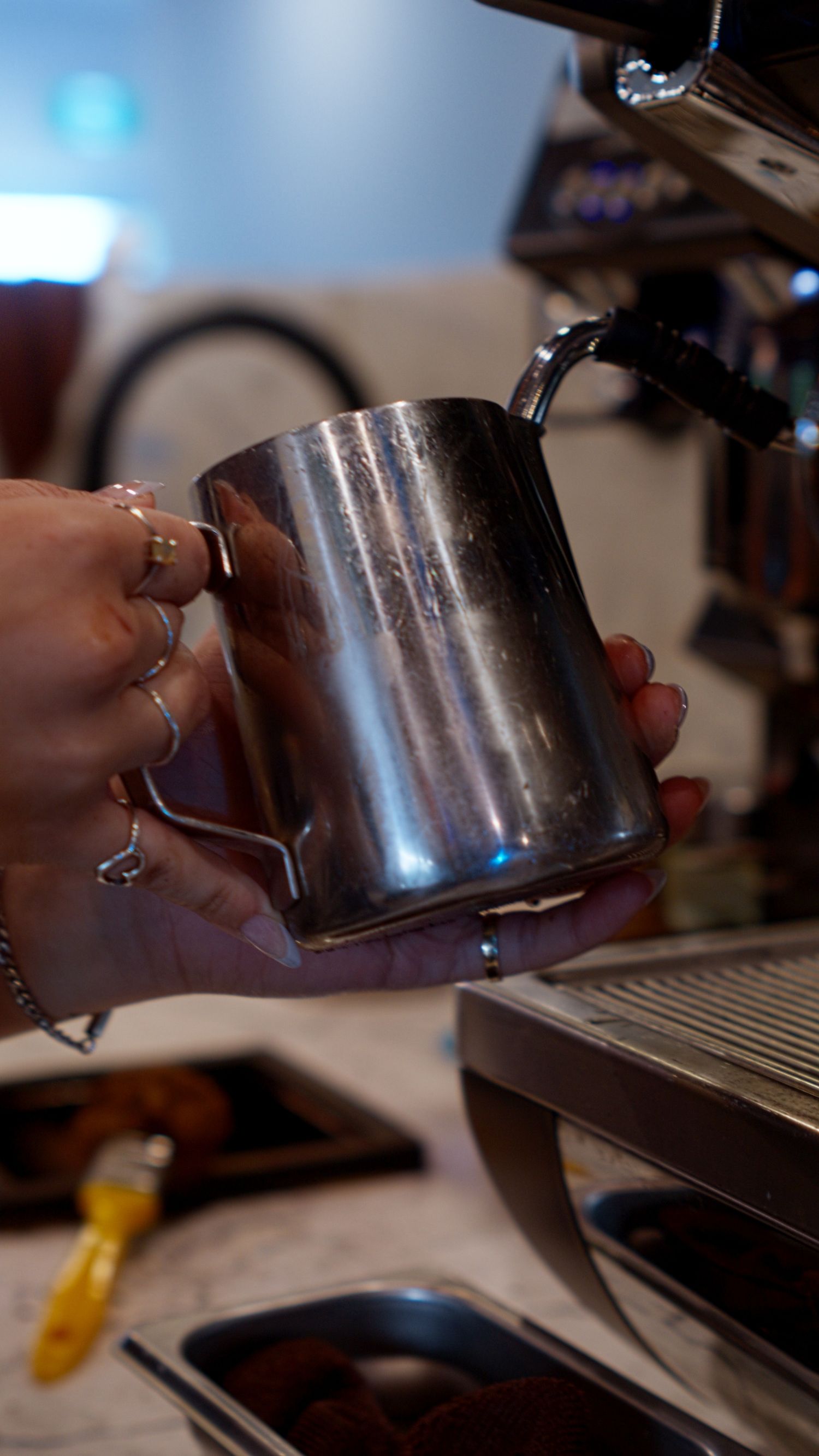 Barista holding shiny stainless steel milk frothing pitcher near professional espresso machine.