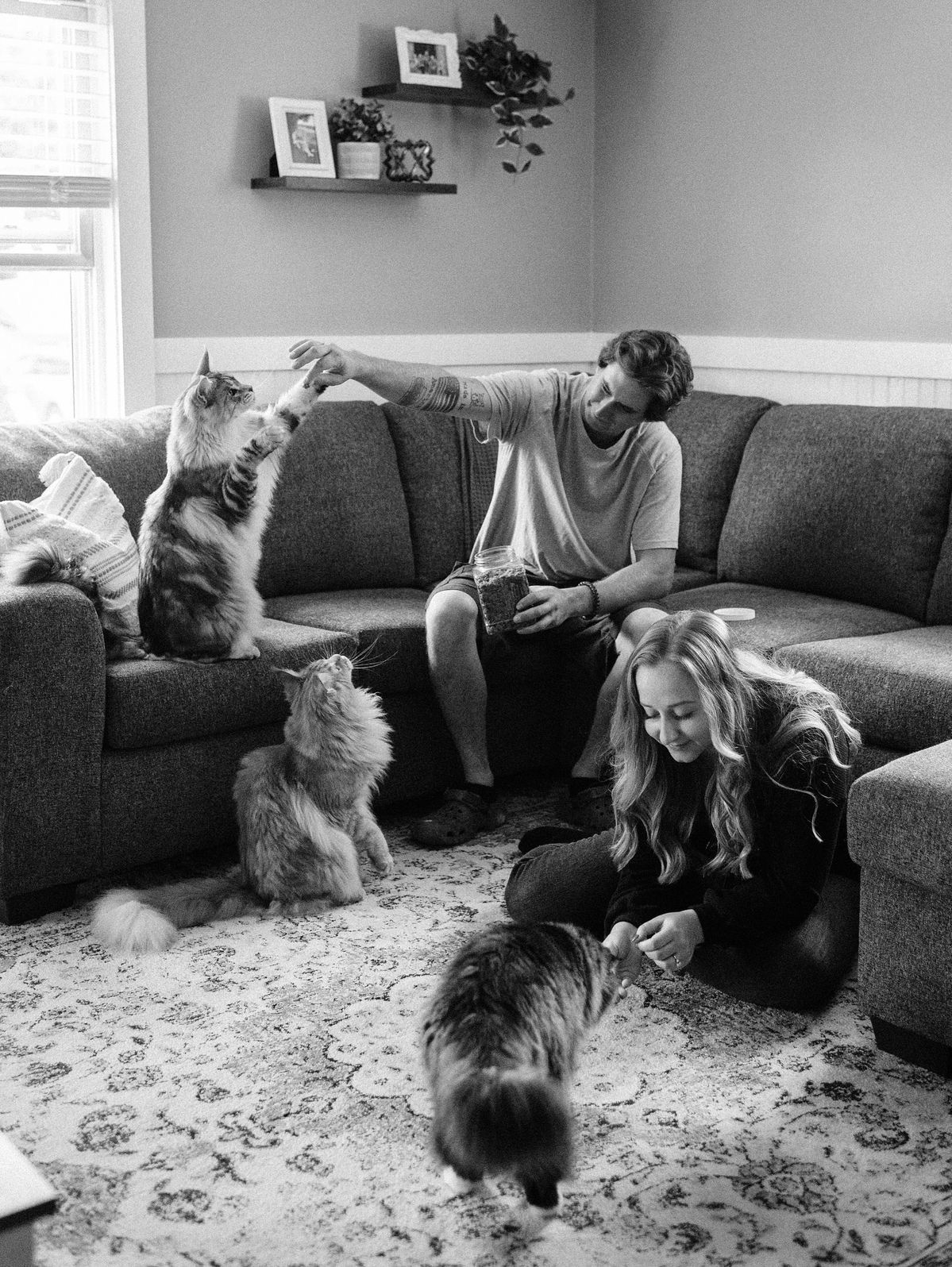 Black and white photo of a couple playing with three fluffy cats in a cozy living room with a sectional sofa.