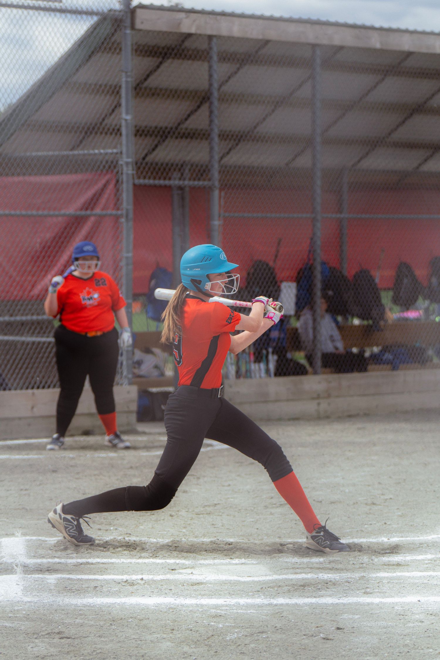 Female softball player in blue helmet and orange jersey swings bat during outdoor game at diamond.