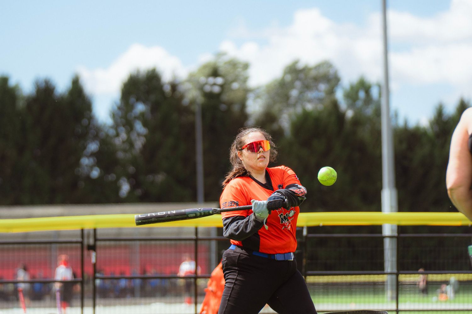 Female softball player in orange uniform swinging bat at yellow ball during outdoor game.