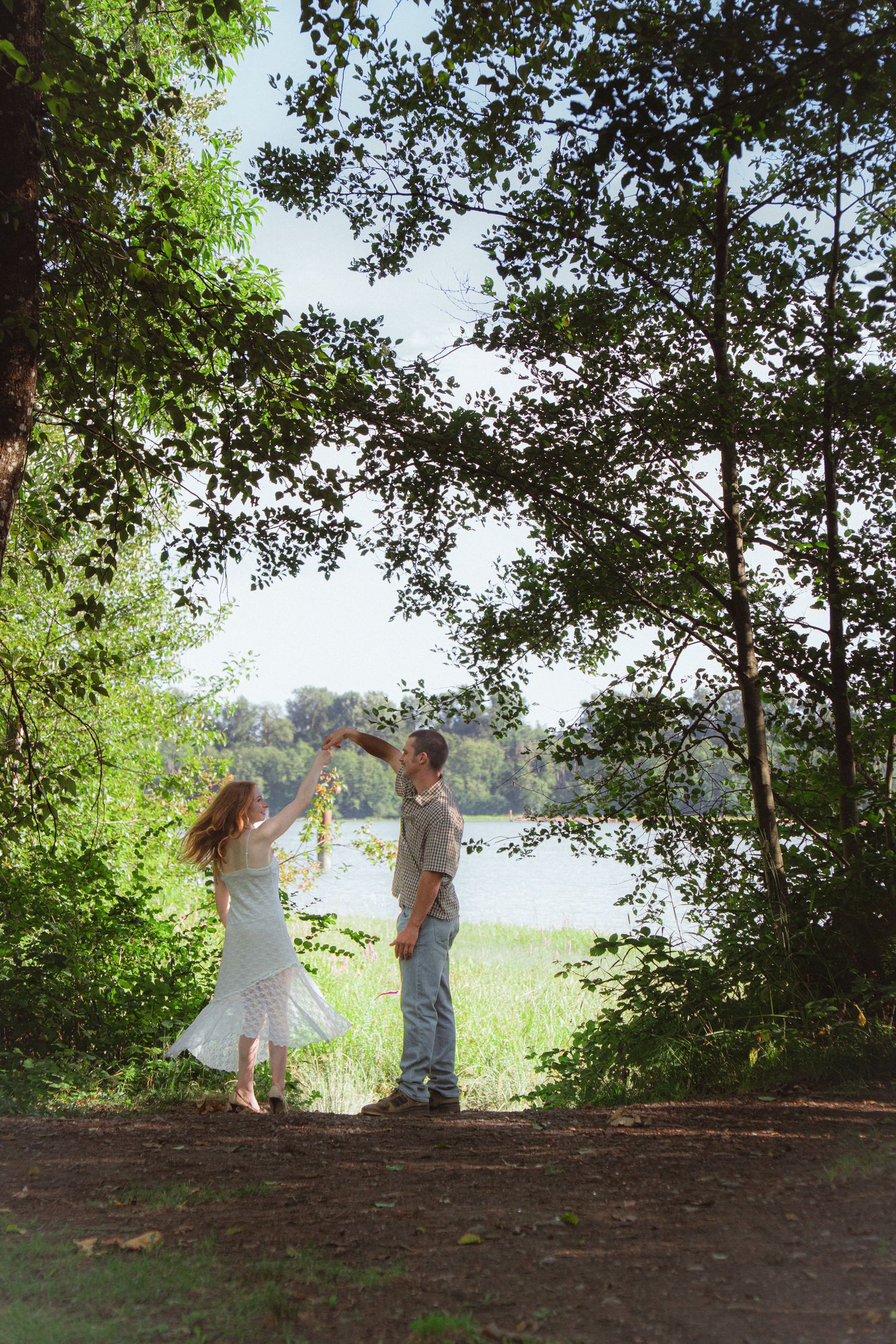 Couple dancing together in a lush green forest near a lake, woman in white dress, man in casual clothes.