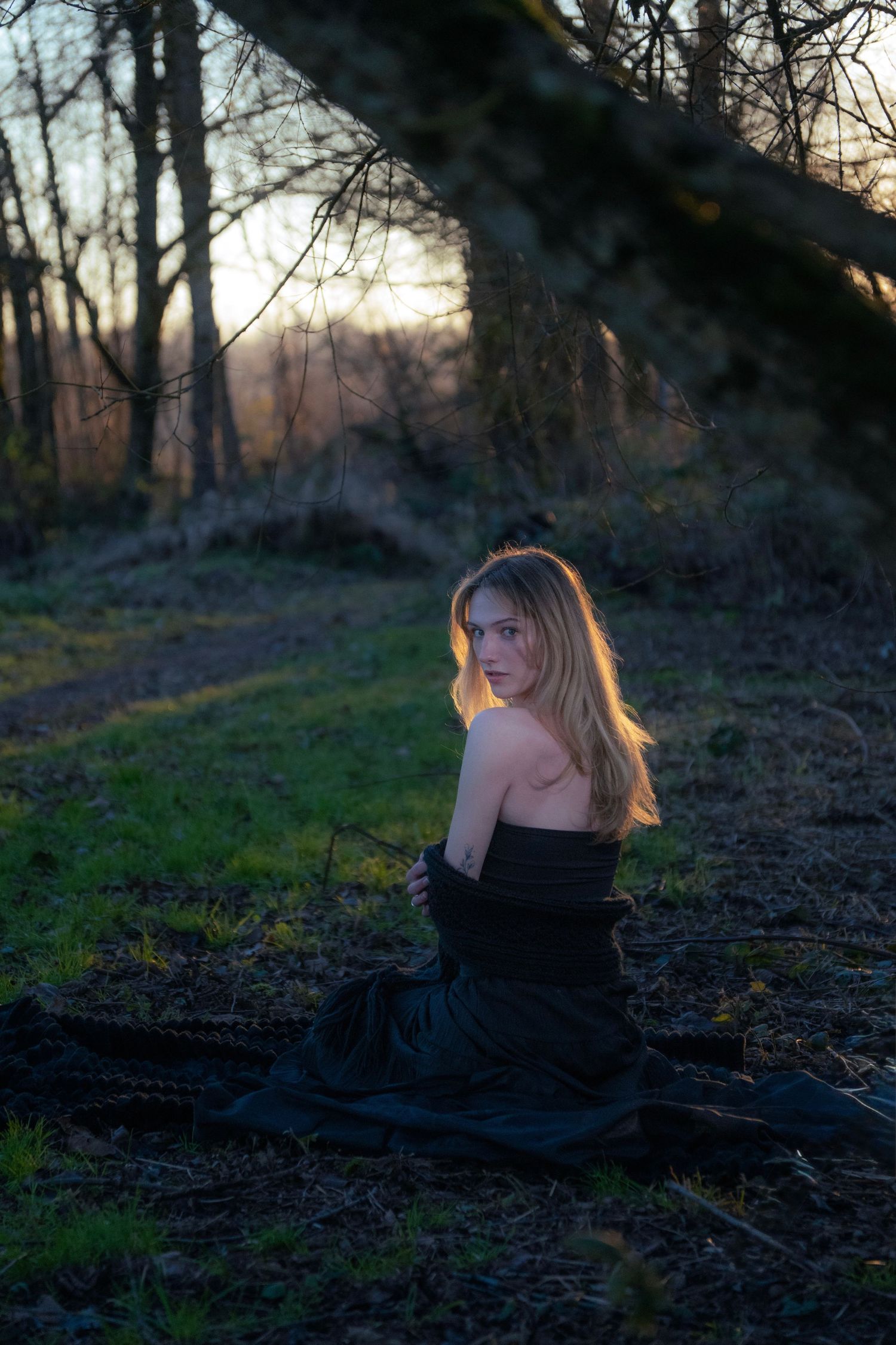 A blonde woman in a black off-shoulder gown poses on forest ground at dusk.