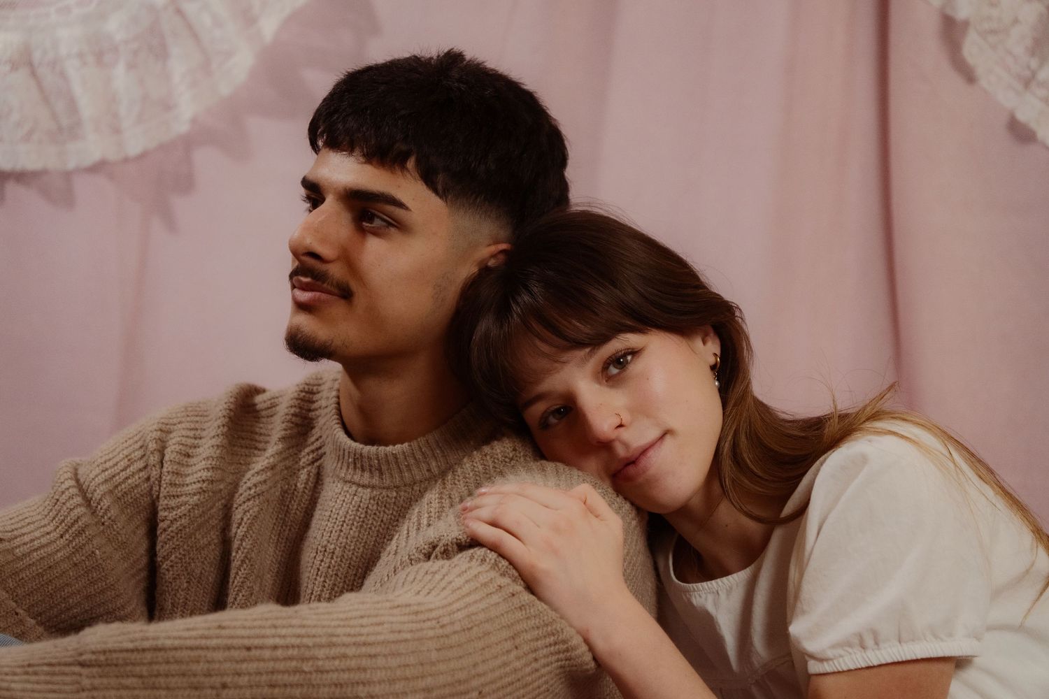 Young couple leaning together romantically against a pink fabric backdrop in a soft portrait.
