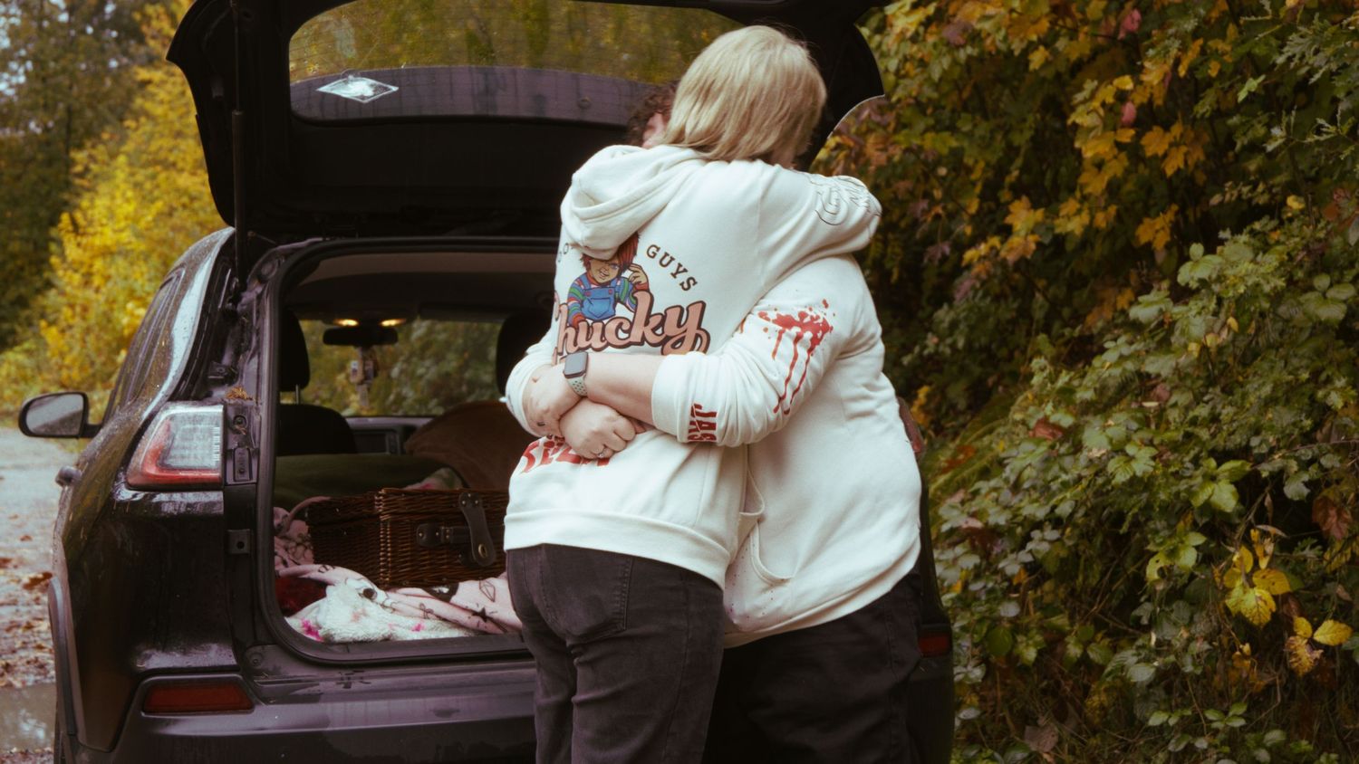 Couple embracing affectionately in matching white graphic hoodies beside an open car trunk surrounded by fall foliage.