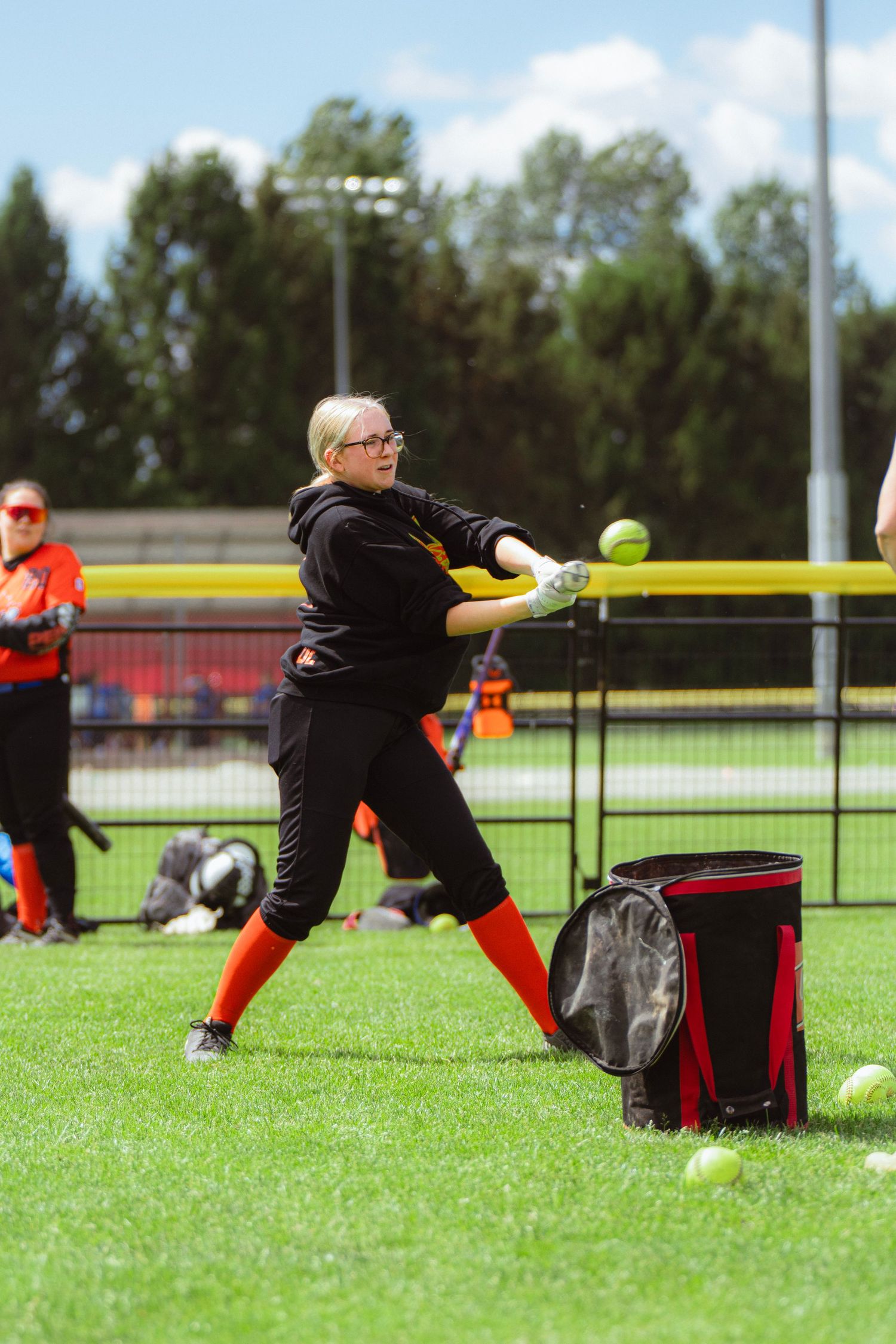 Blonde female softball player hitting ball near equipment bag on field.