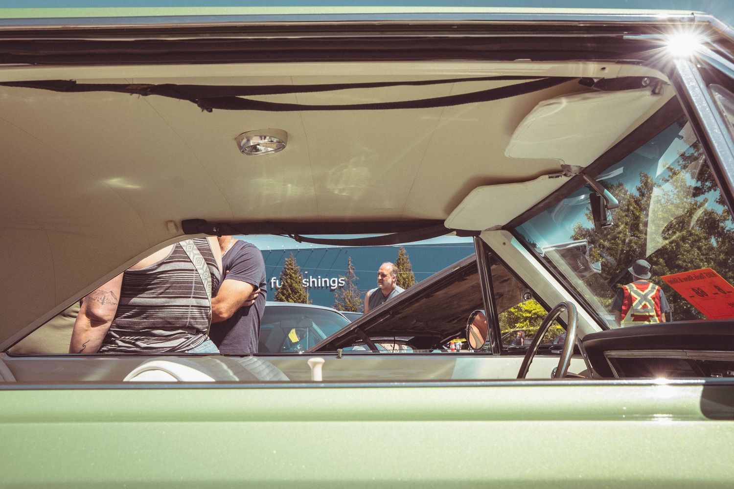 View through vintage green car window reflecting people at an outdoor car show event.