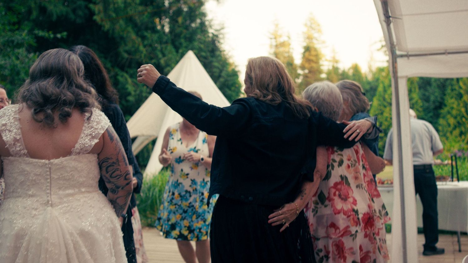 A bride in white leads a group dance with guests in colorful dresses at an outdoor wedding reception.