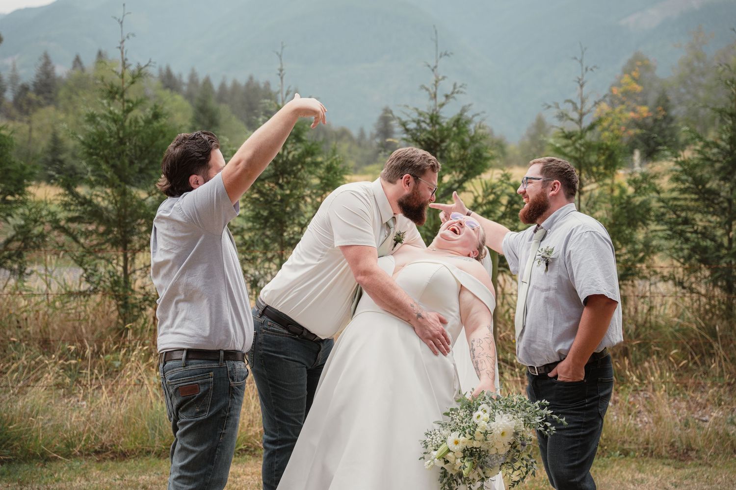 Groom dips bride for kiss while groomsmen celebrate at outdoor mountain wedding.