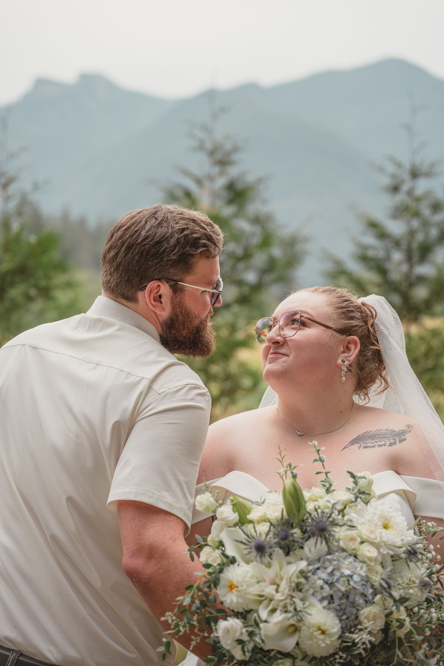 Bride and groom share a loving glance outdoors, with mountain views and evergreen trees, bride holding white floral bouquet.