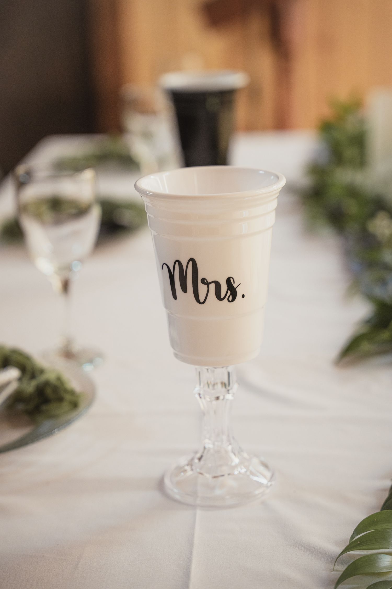 White ceramic Mrs. cup on crystal stem goblet at elegant wedding reception table with greenery.