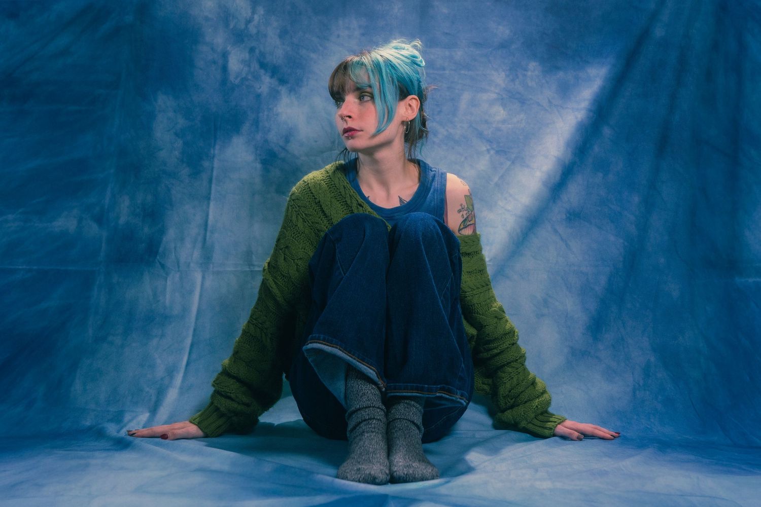 Young woman with teal hair wearing green cardigan and blue dress sits on studio floor against blue backdrop.