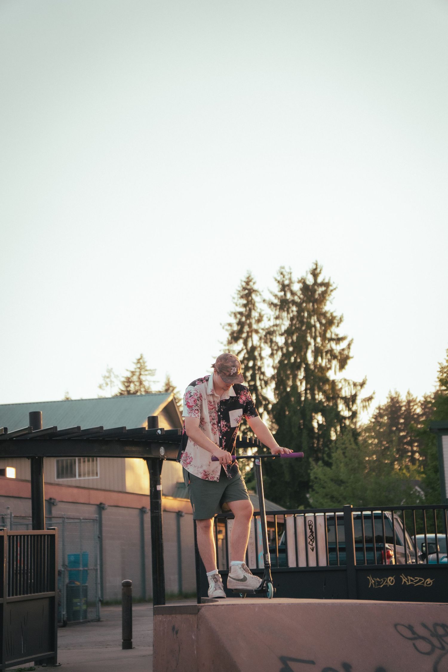 Man in floral shirt standing on a scooter atop a ramp at a skate park during golden hour.