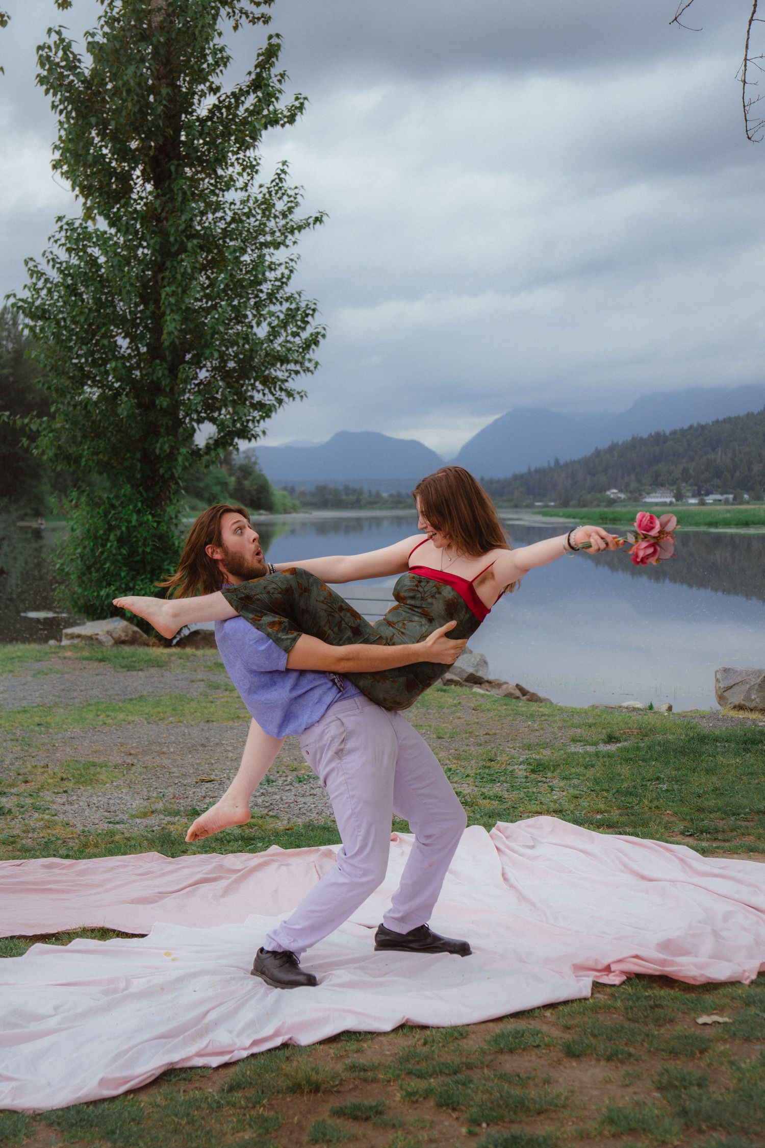 Man and woman dance together on pink blanket by a scenic mountain lake on cloudy day.