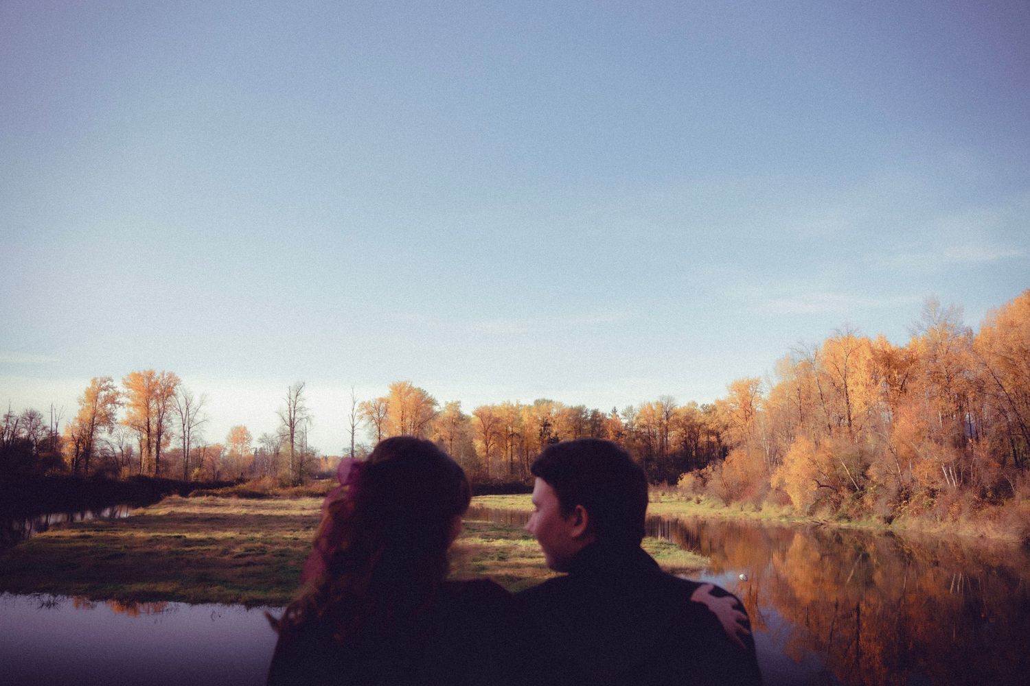Couple sitting together by a calm river surrounded by autumn trees with golden foliage under a clear blue sky.