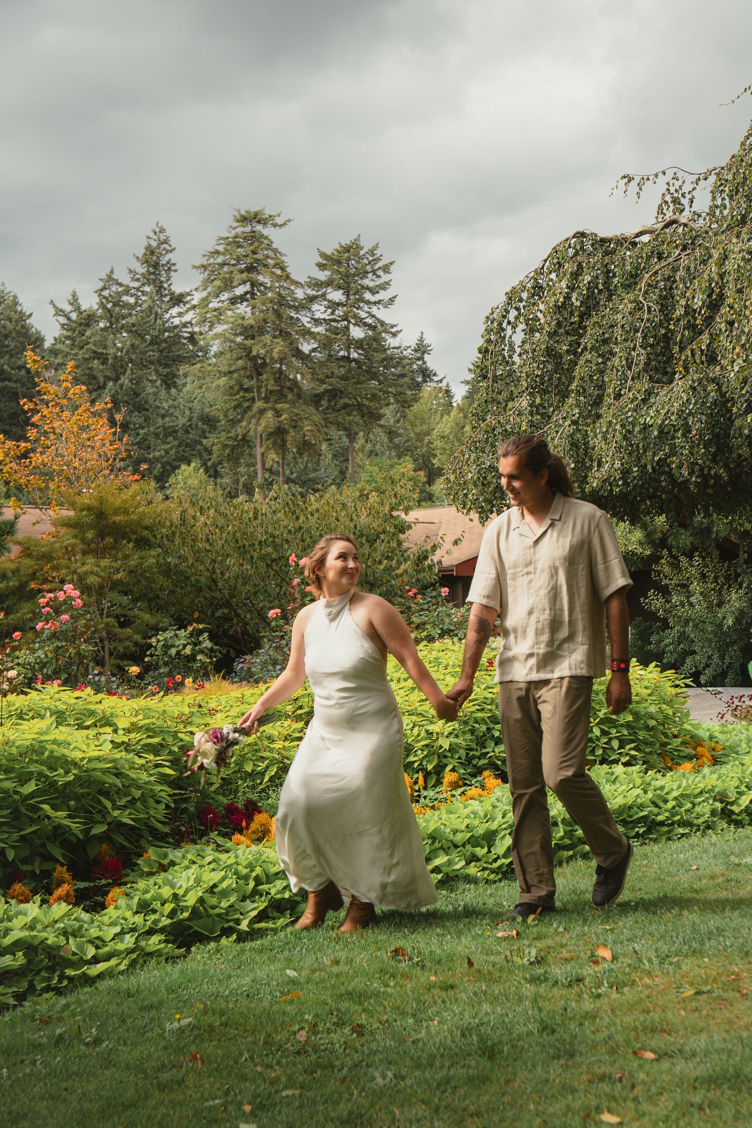 Newlyweds walk hand-in-hand through a vibrant garden with colorful blooms and tall evergreen trees.