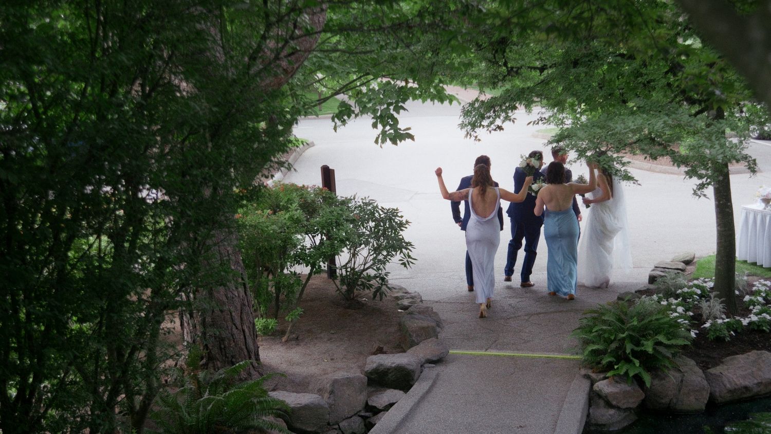Group of people walking on a stone path through lush green trees toward a white building outdoors.